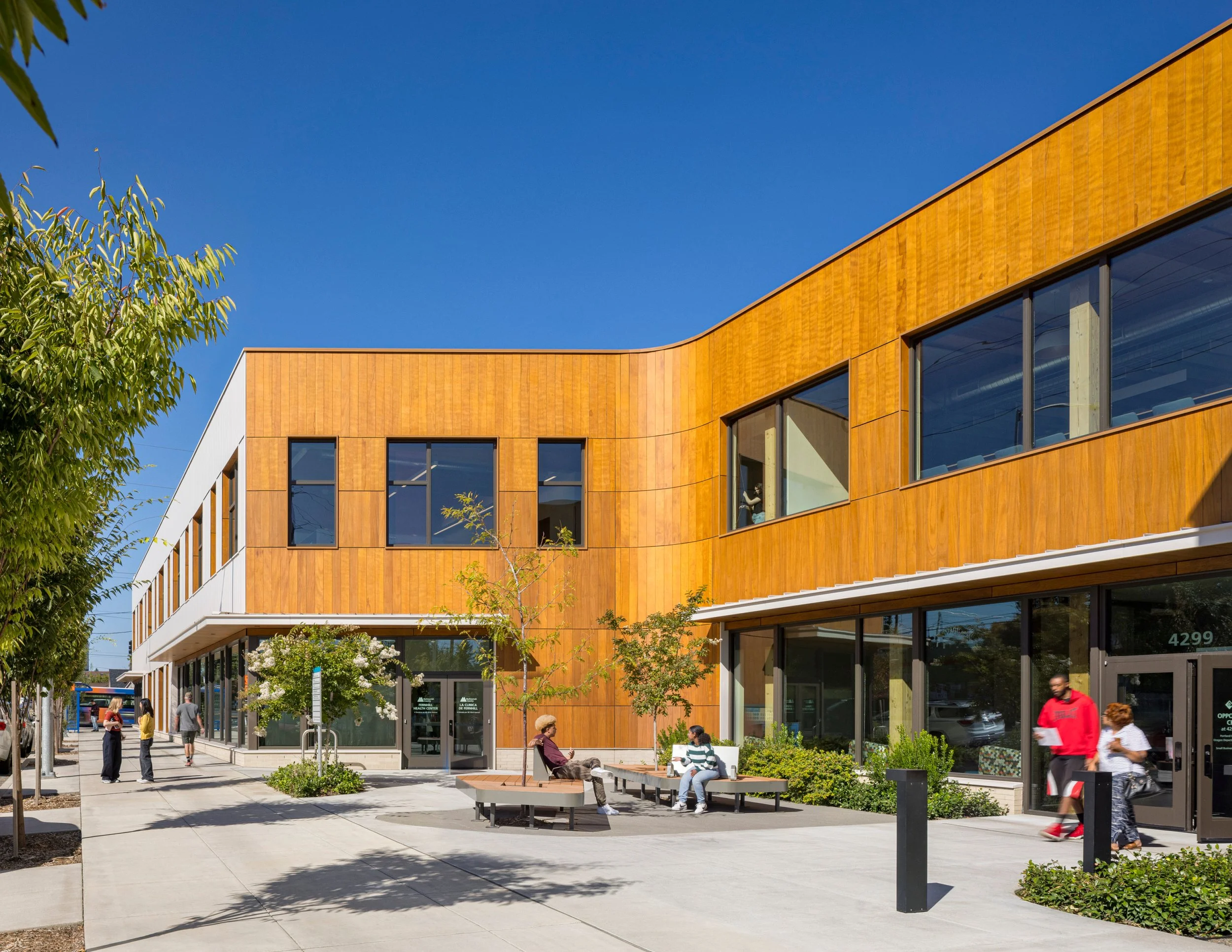 Exterior of a modern elementary school with a large wooden overhang and multiple windows, students and adults outside on a sunny day with blue skies and some clouds.