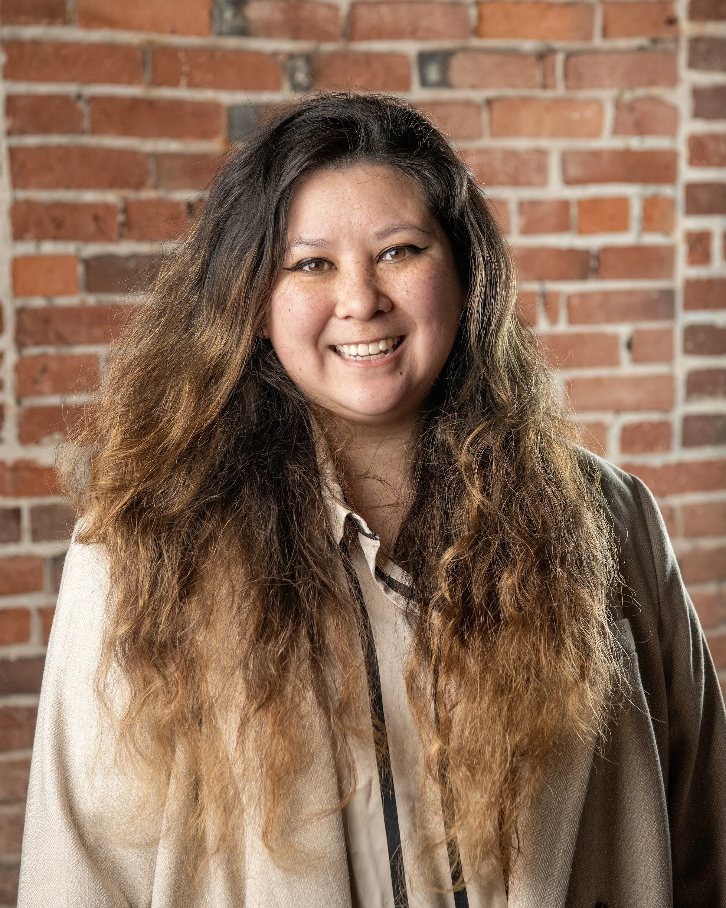 A young woman with long, wavy brown hair, smiling and wearing a black shirt, standing indoors against a neutral background.