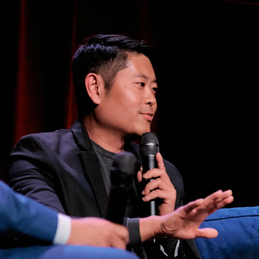man speaking into a microphone during a discussion or interview, seated on a stage with a dark background.