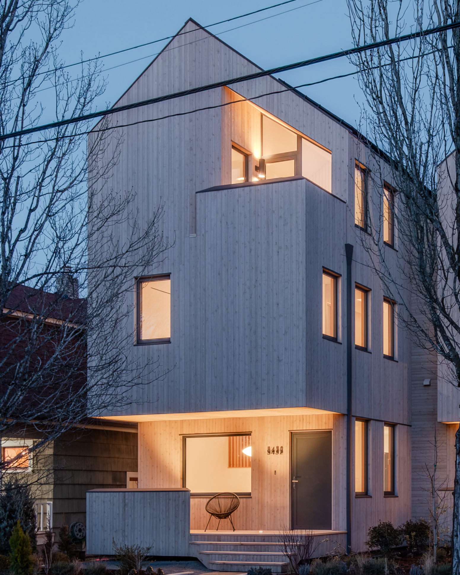 Modern multi-story house with a wooden facade, large glass windows, and a small garden area in front, shadow of a tree on the building.