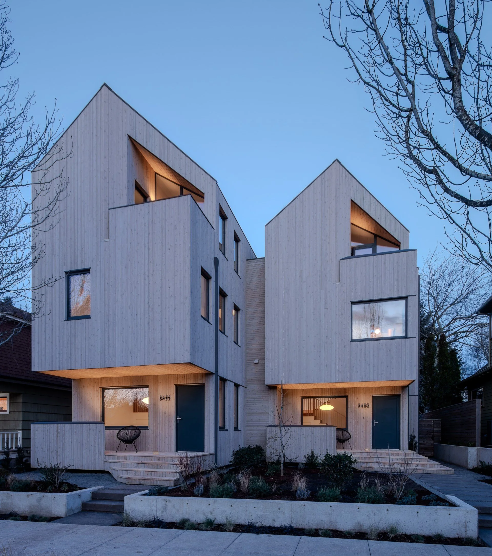 Modern multi-story house with a wooden facade, large glass windows, and a small garden area in front, shadow of a tree on the building.