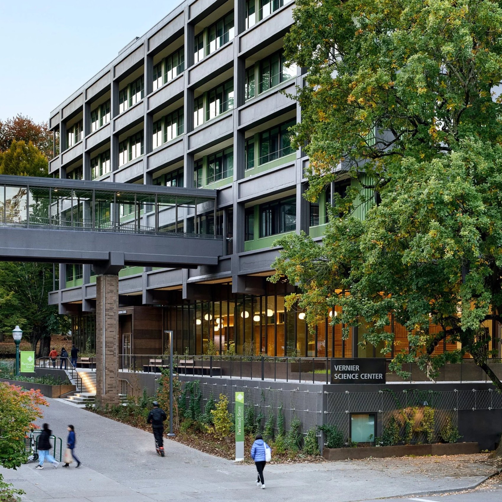 Modern building with large windows and solar panels on the roof, street lamp, and small tree in front, during dusk.