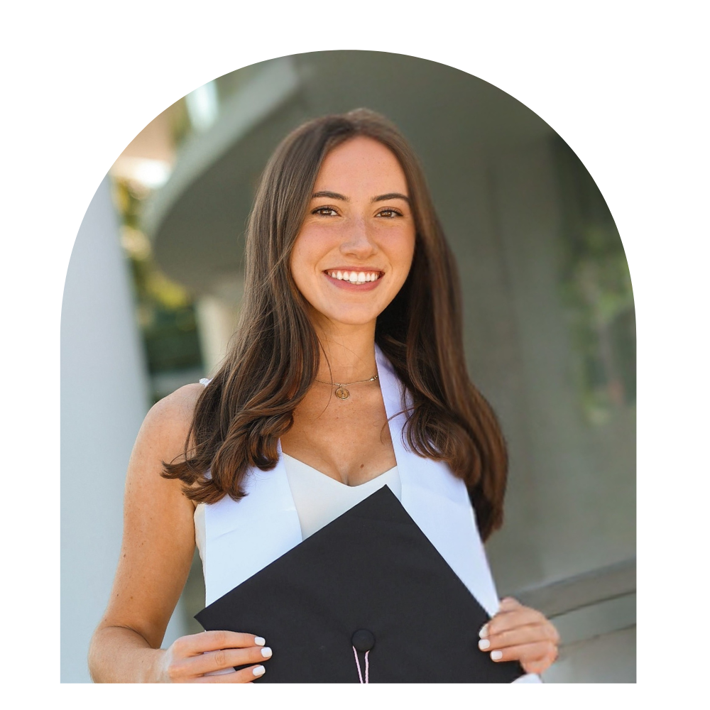 A young woman with long brown hair smiling while holding a graduation cap, wearing a white top, outdoors in daylight.