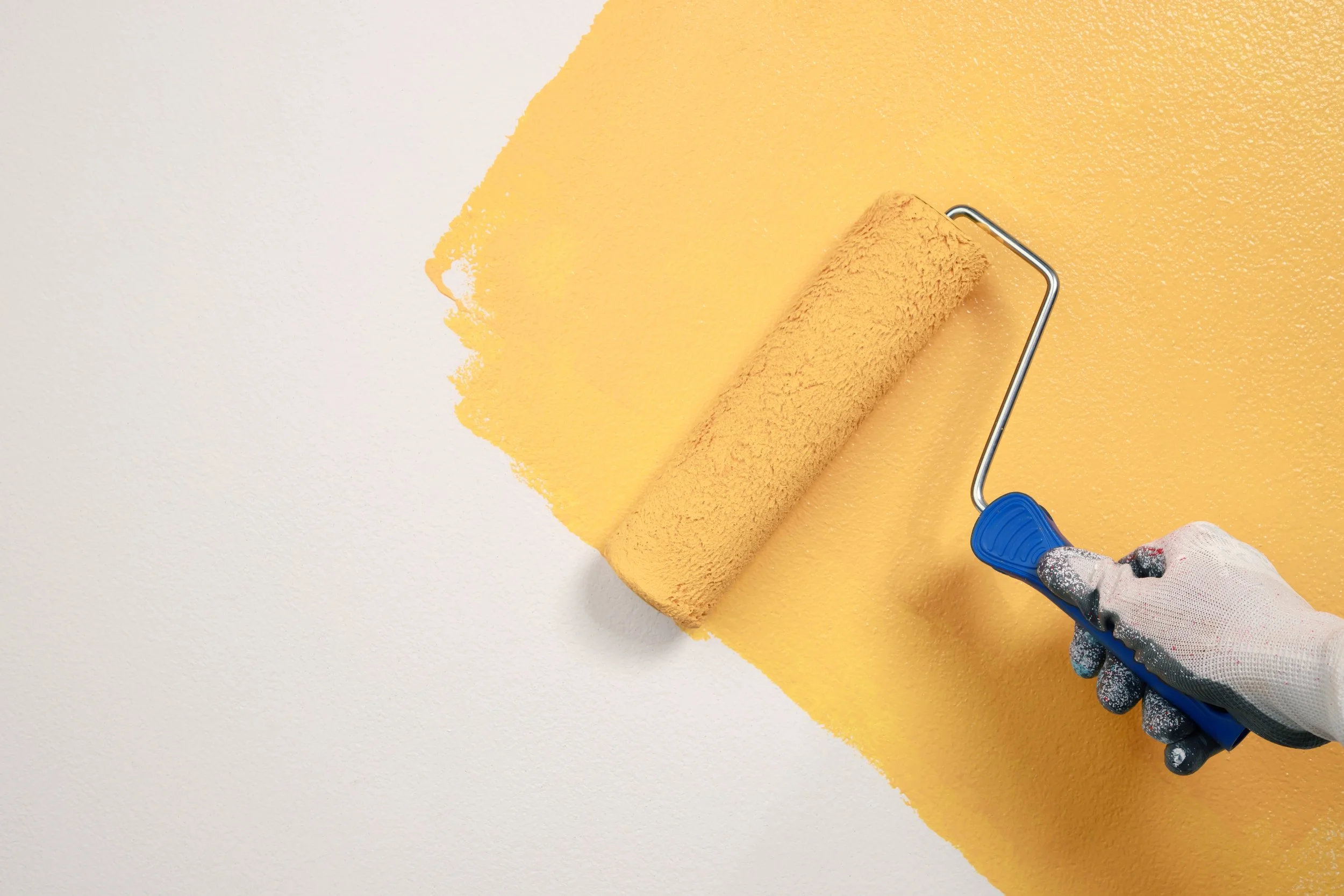 Person painting a wall with a paint roller, applying yellow paint over white wall.