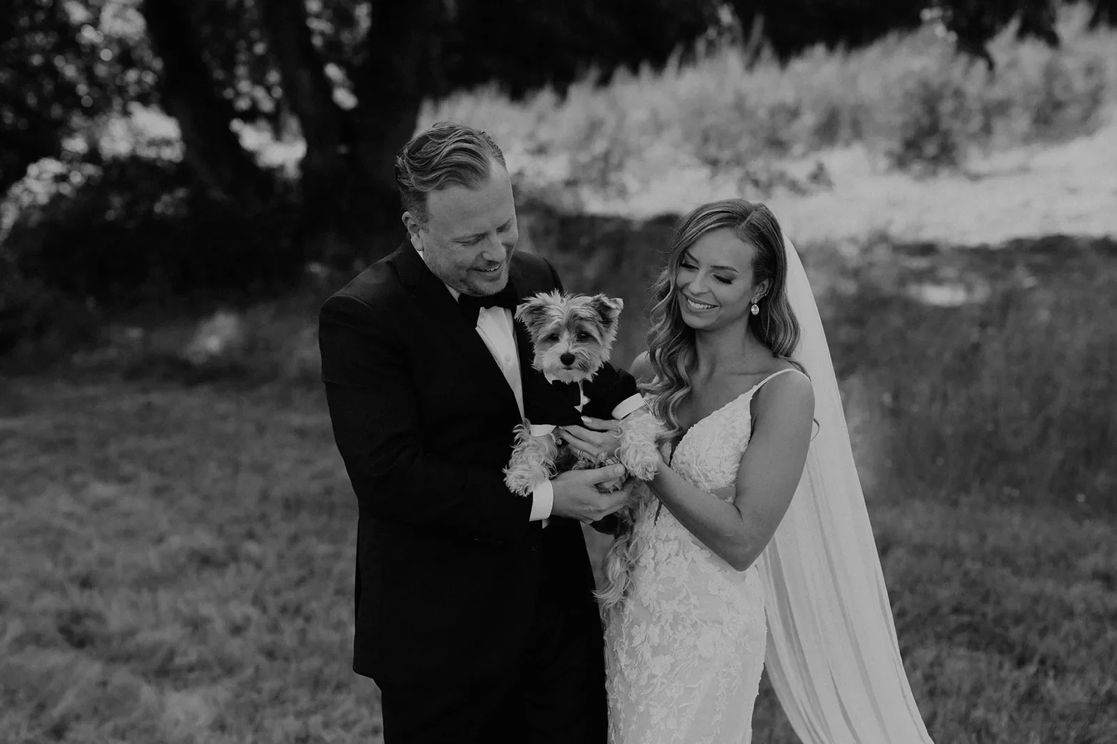Bride and groom holding a small dog at their wedding outdoors, smiling and dressed in wedding attire, with trees in the background.