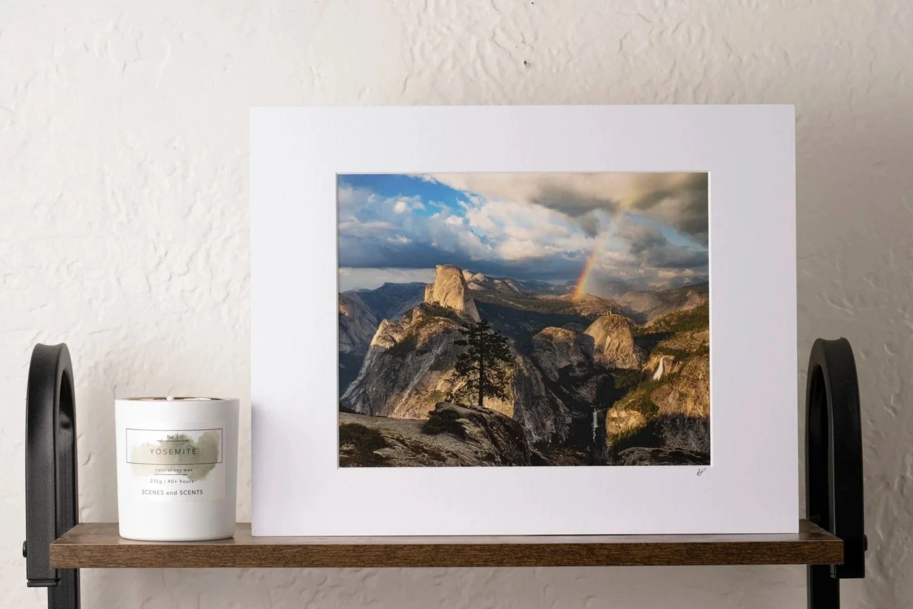 Framed landscape photograph of Yosemite National Park showing granite cliffs, trees, a rainbow, and a waterfall in the distance on a wooden shelf, with a white scented candle labeled 'Yosemite' next to it.