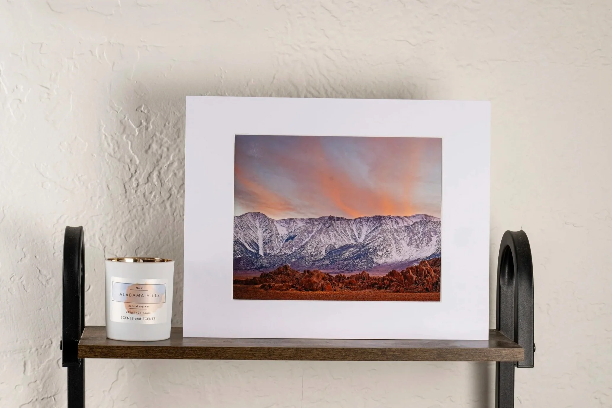 Photo of a framed landscape picture of snow-capped mountains and a colorful sky, placed on a wooden and metal shelf. To the left of the frame is a white candle with a gold rimmed lid labeled 'Alabama Hills'.