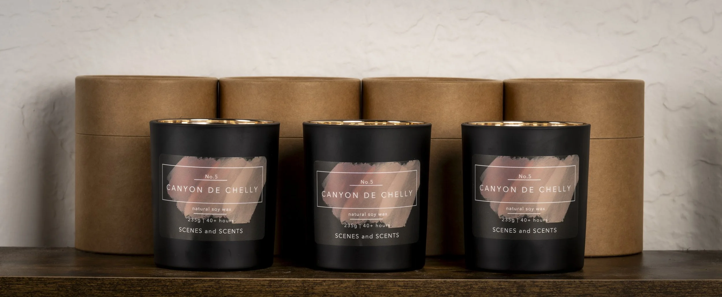 Three black candles with labels that read 'Canyon de Chelly' and 'natural soy wax', placed in front of a round, brown cardboard box. The candles are on a wooden surface against a textured white wall.