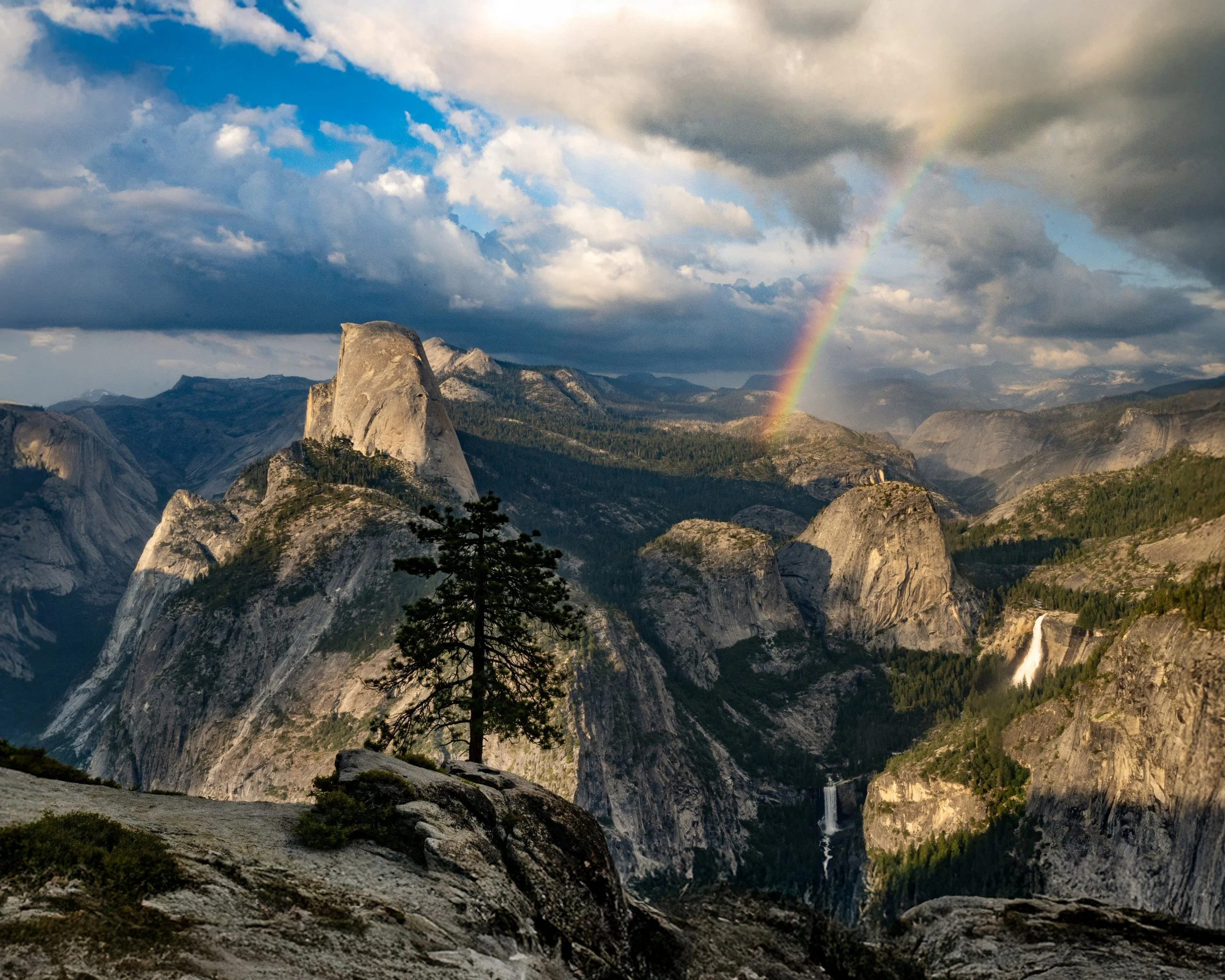 Scenic view of Yosemite National Park showing granite cliffs, waterfalls, trees, a rainbow, and a cloudy sky.