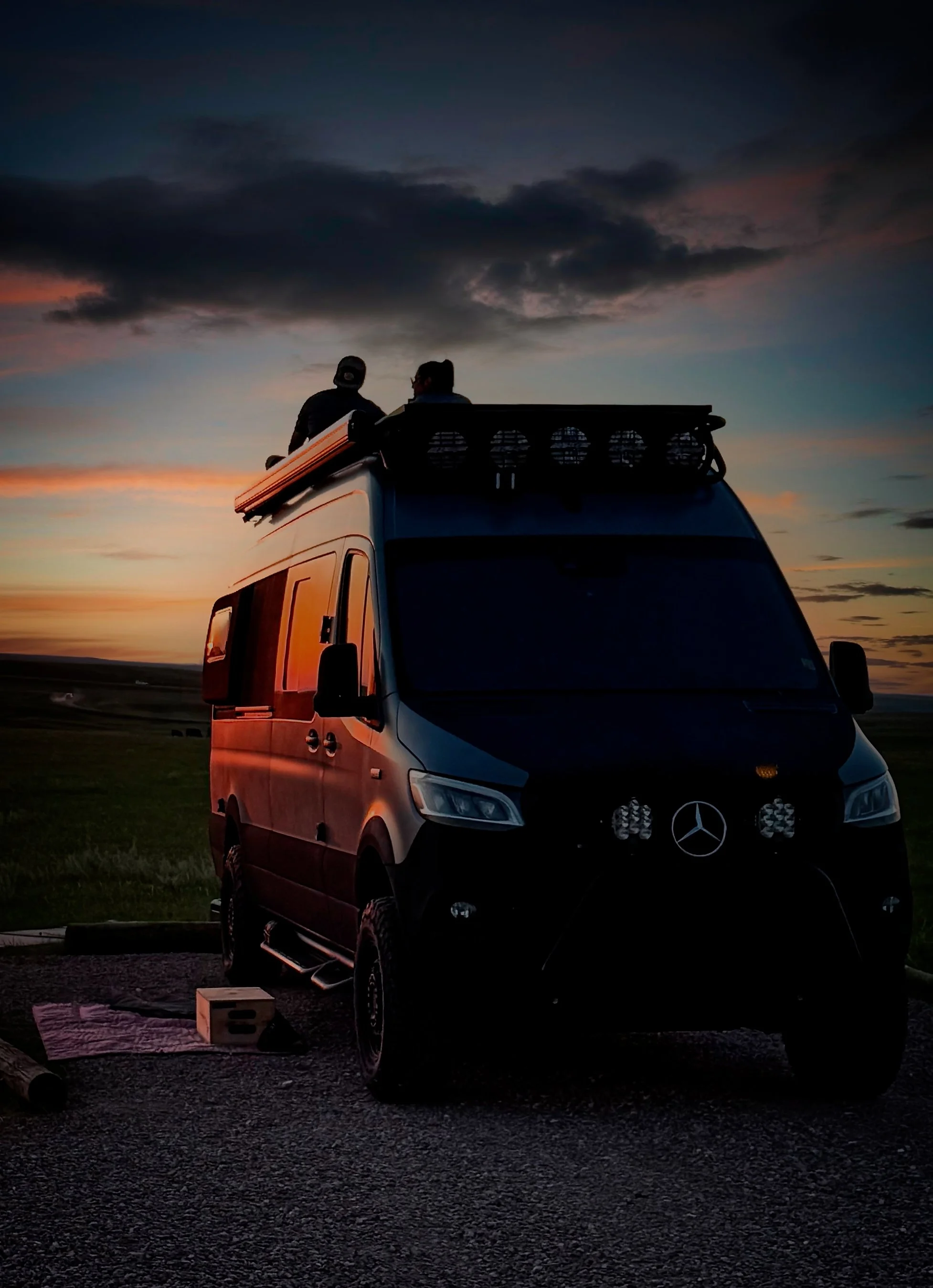 A black van with roof lights parked on an open field during sunset, with two people sitting on top of the van, watching the sky.