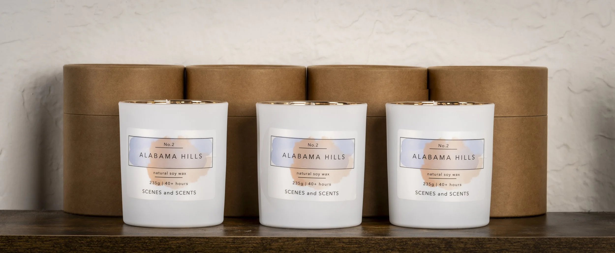 Three white candles labeled 'Alabama Hills' placed in front of three brown cylindrical boxes on a wooden surface.