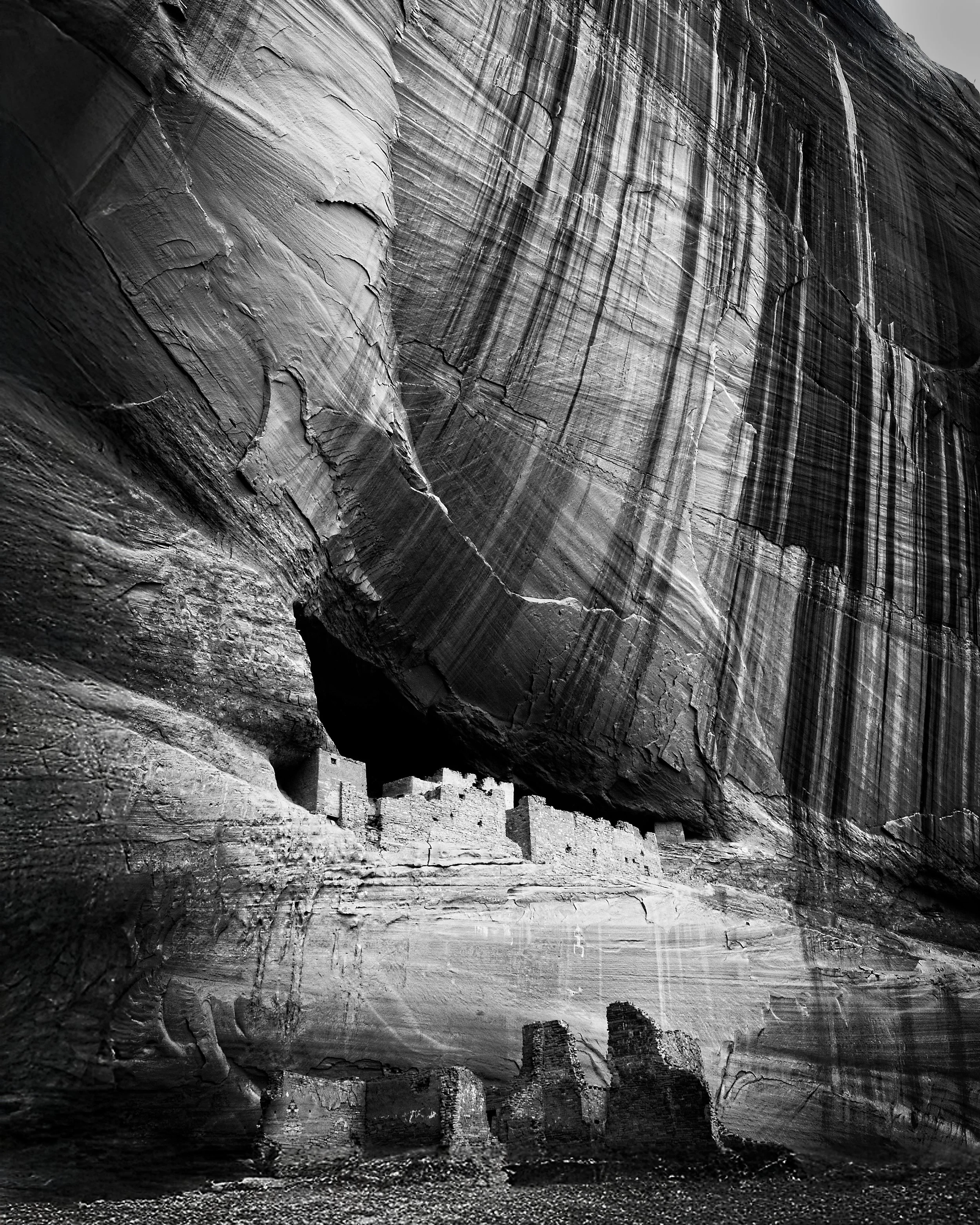 Ancient cliff dwelling built into a massive rock face with layered, striated patterns, in black and white.