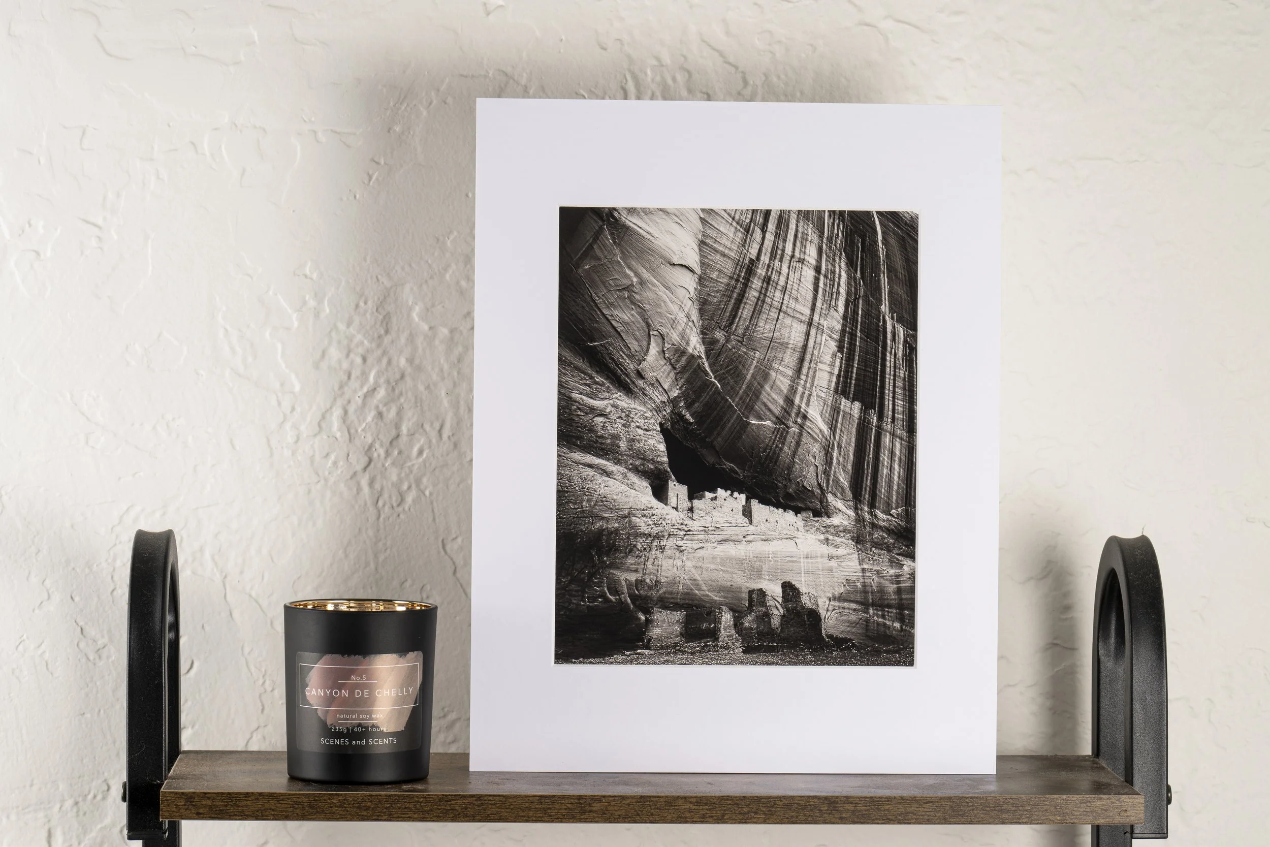 Black and white photograph of a canyon with rock formations, displayed on a white board or mat, placed on a wooden shelf against a textured white wall. A black candle with a label reading 'Canyon de Chelly' is on the left side of the shelf.