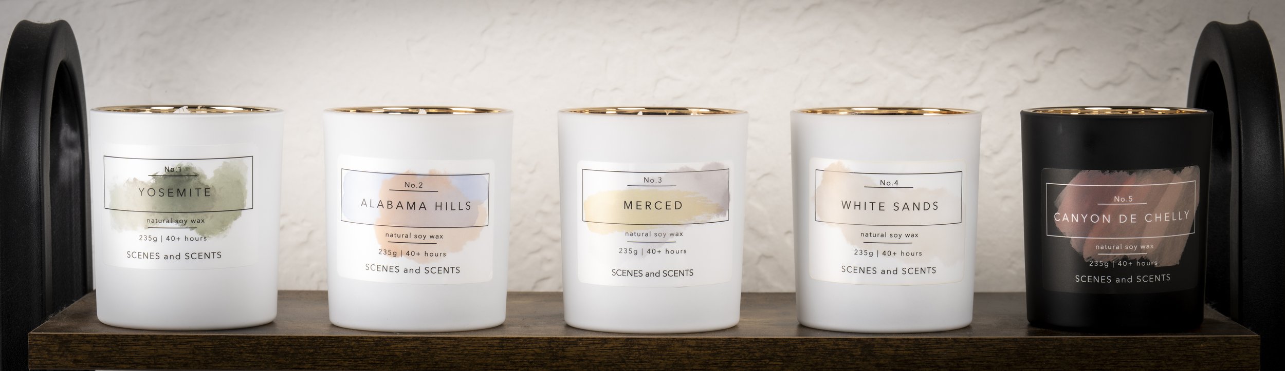 Five candles with different labels arranged on a wooden shelf. The candles are labeled Yosemite, Alabama Hills, Merced, White Sands, and Canyon De Chelly.