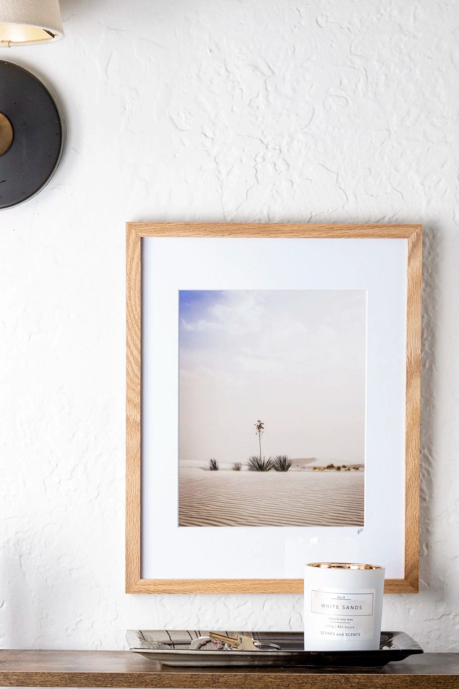A framed photograph of a desert landscape with a solitary tree and sand dunes, hung on a textured white wall. A small candle labeled 'White Sands' is on a tray in front of the frame.
