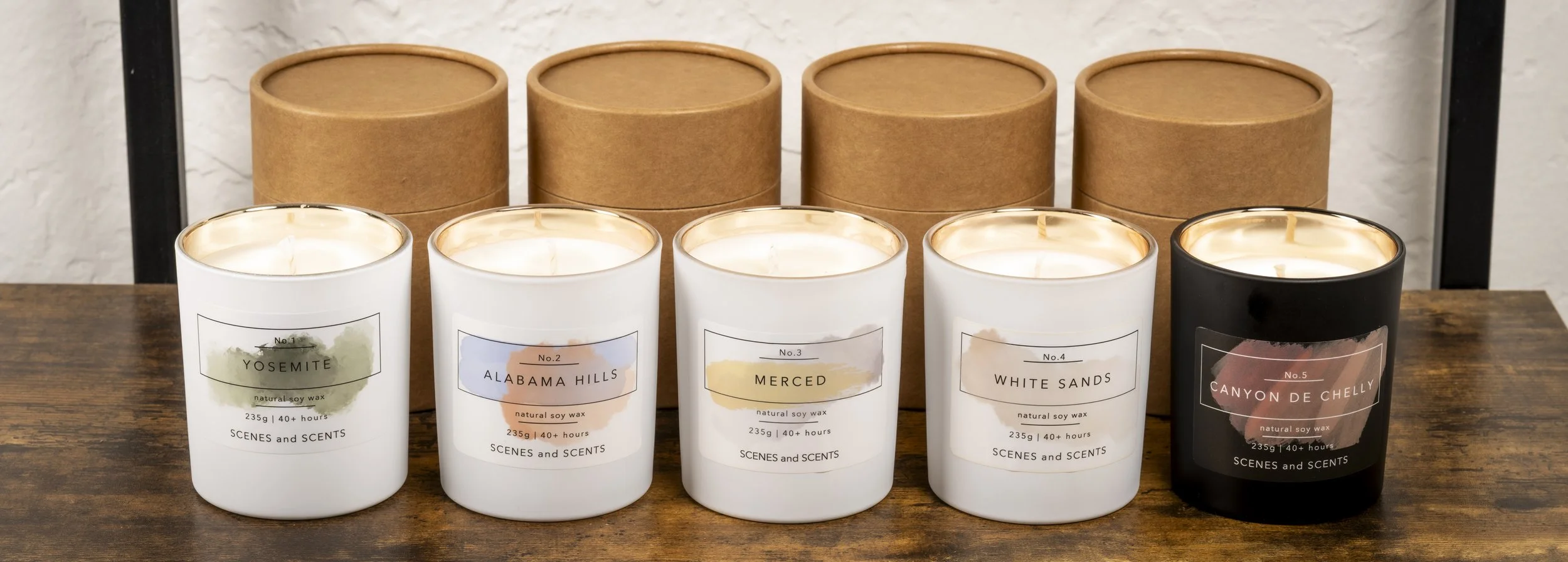 Five scented candles in white and black containers labeled Yosemite, Alabama Hills, Merced, White Sands, and Canyon de Chelly, arranged on a wooden surface with brown boxes in the background.