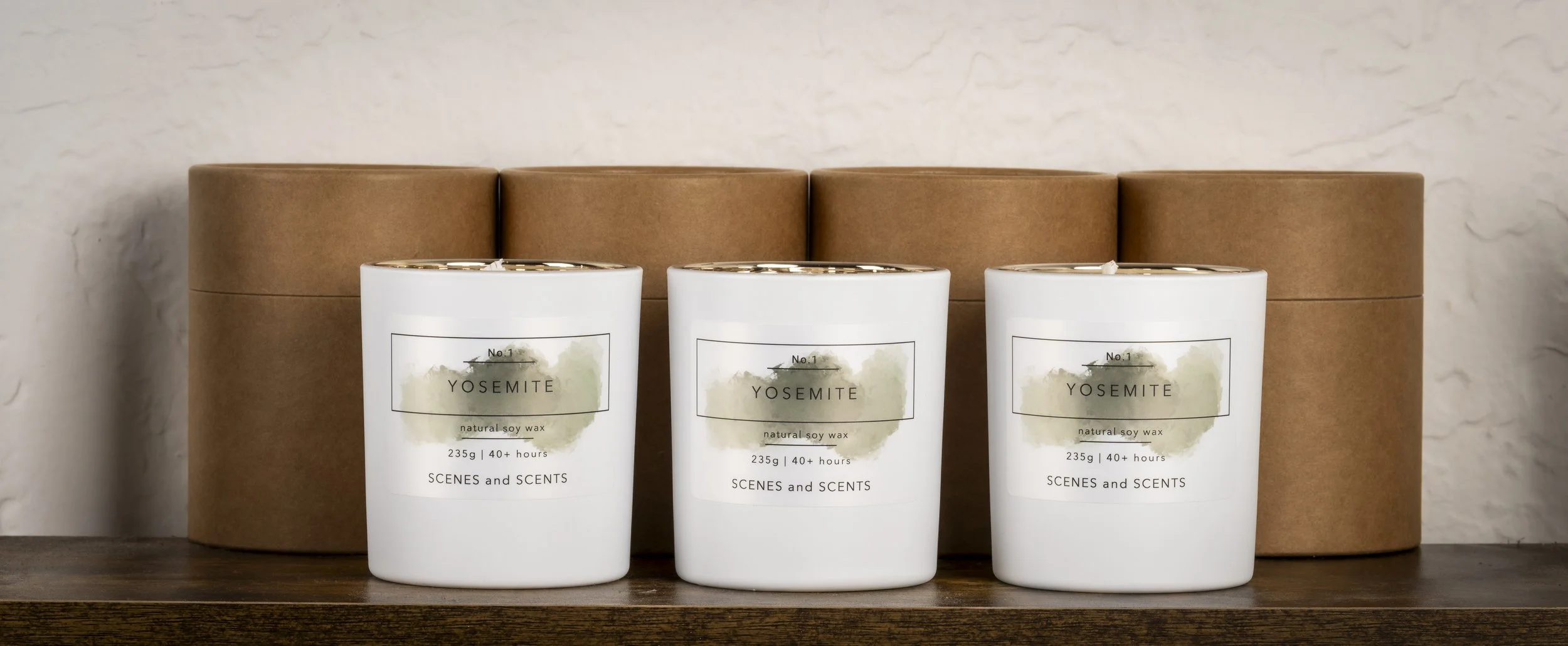 Three white candles labeled Yosemite natural soy wax with 40+ hours burn time, placed on a wooden surface in front of three round, brown cardboard storage boxes against a textured wall.