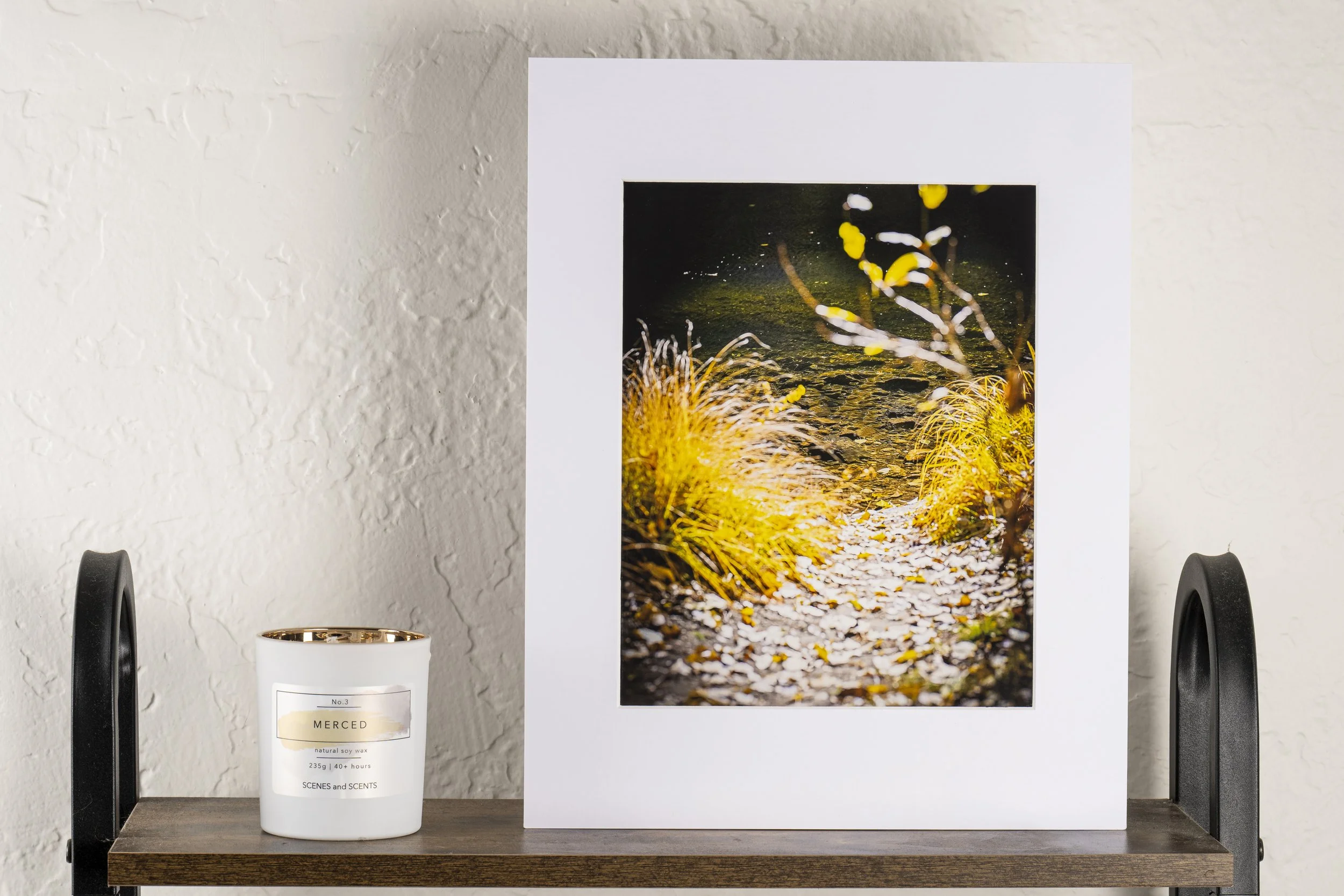 A framed photograph of a nature scene with yellow grasses, a pathway covered in leaves, and a dark water body in the background, placed on a wooden shelf with a white candle next to it.