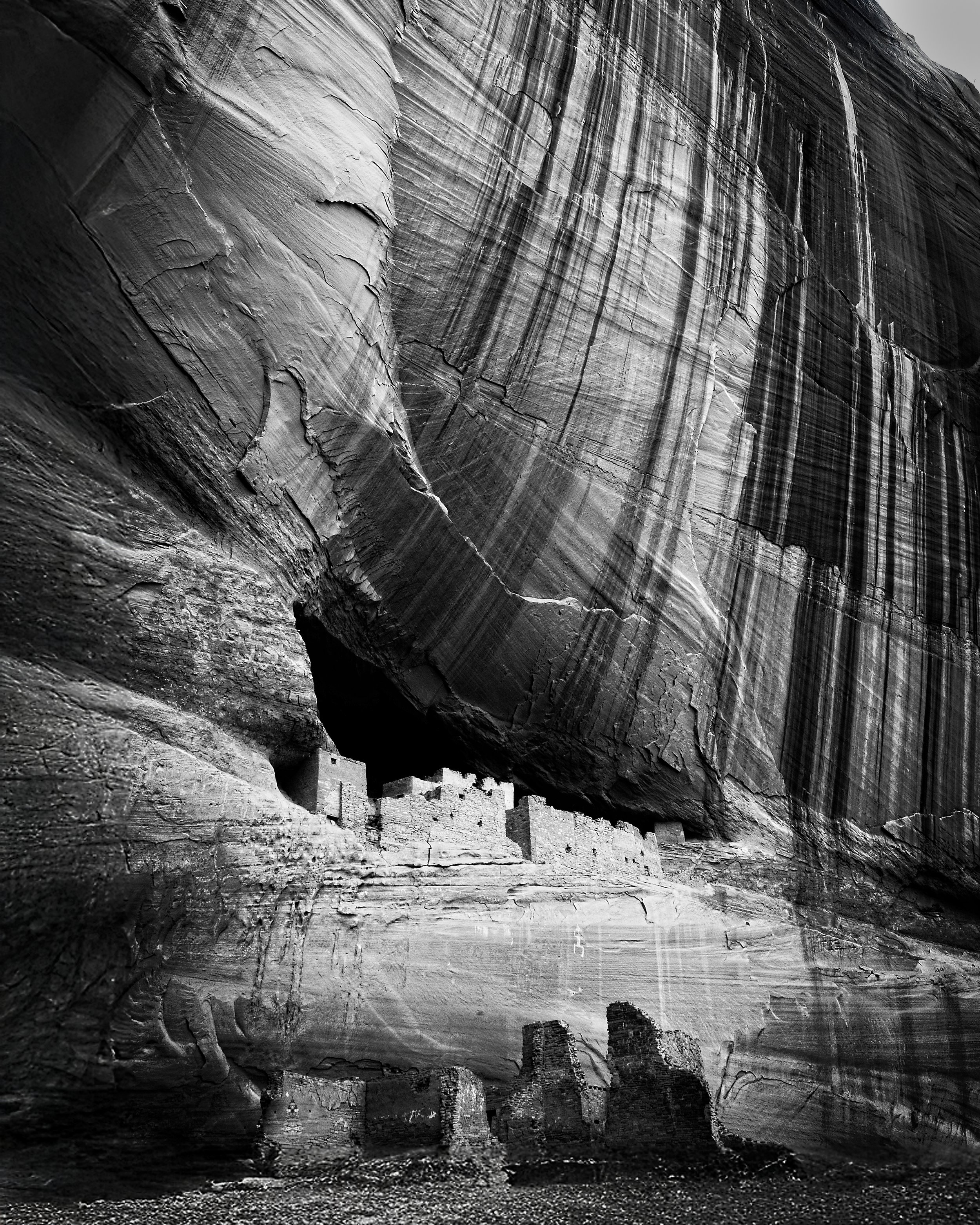 Ancient cliff dwellings built into the rock face of a tall canyon wall with vertical striations in a black and white photograph.