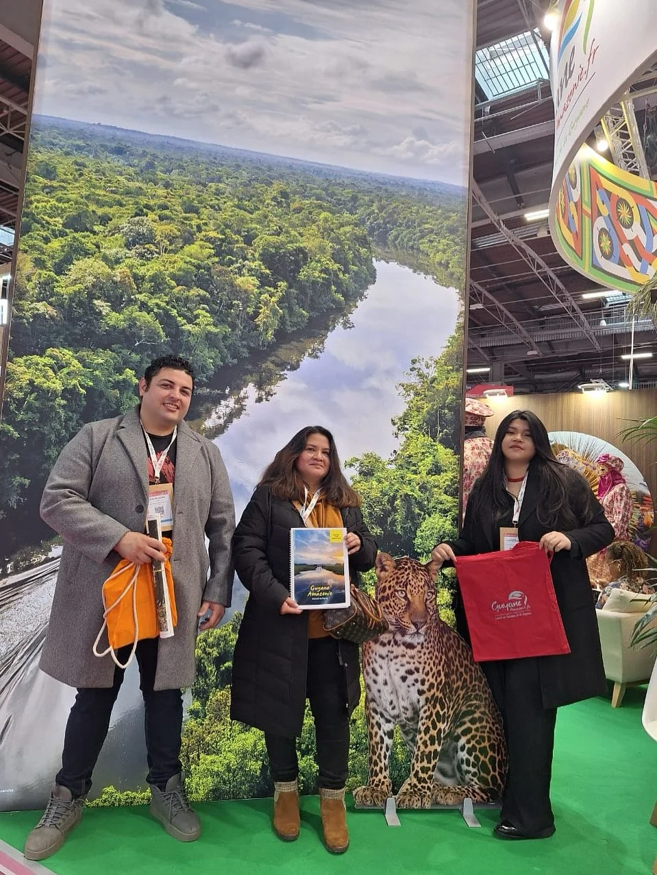 Trois personnes posent devant une grande image de la forêt amazonienne avec un fleuve, une femme tient un livre sur la Guyane, une autre femme tient une affiche avec un jaguar en plastique.