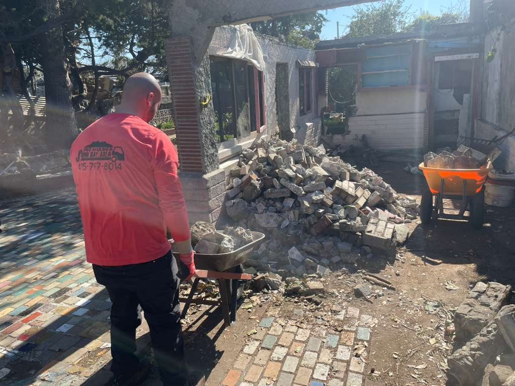 A person working with a wheelbarrow and pile of rubble in a damaged outdoor area with a partially destroyed building.