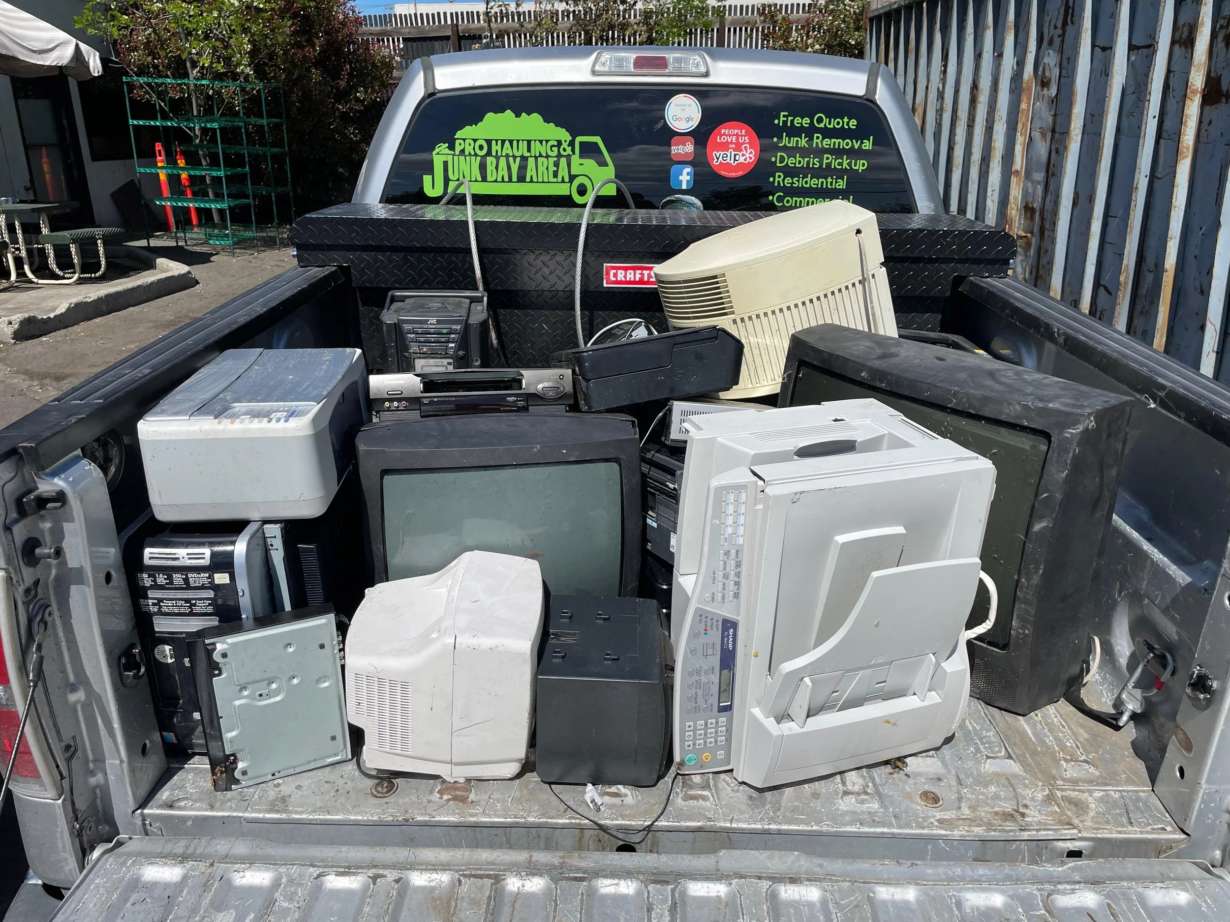 Pickup truck bed filled with old electronics including CRT monitors, printers, and other electronic devices, parked outdoors next to a wooden fence.