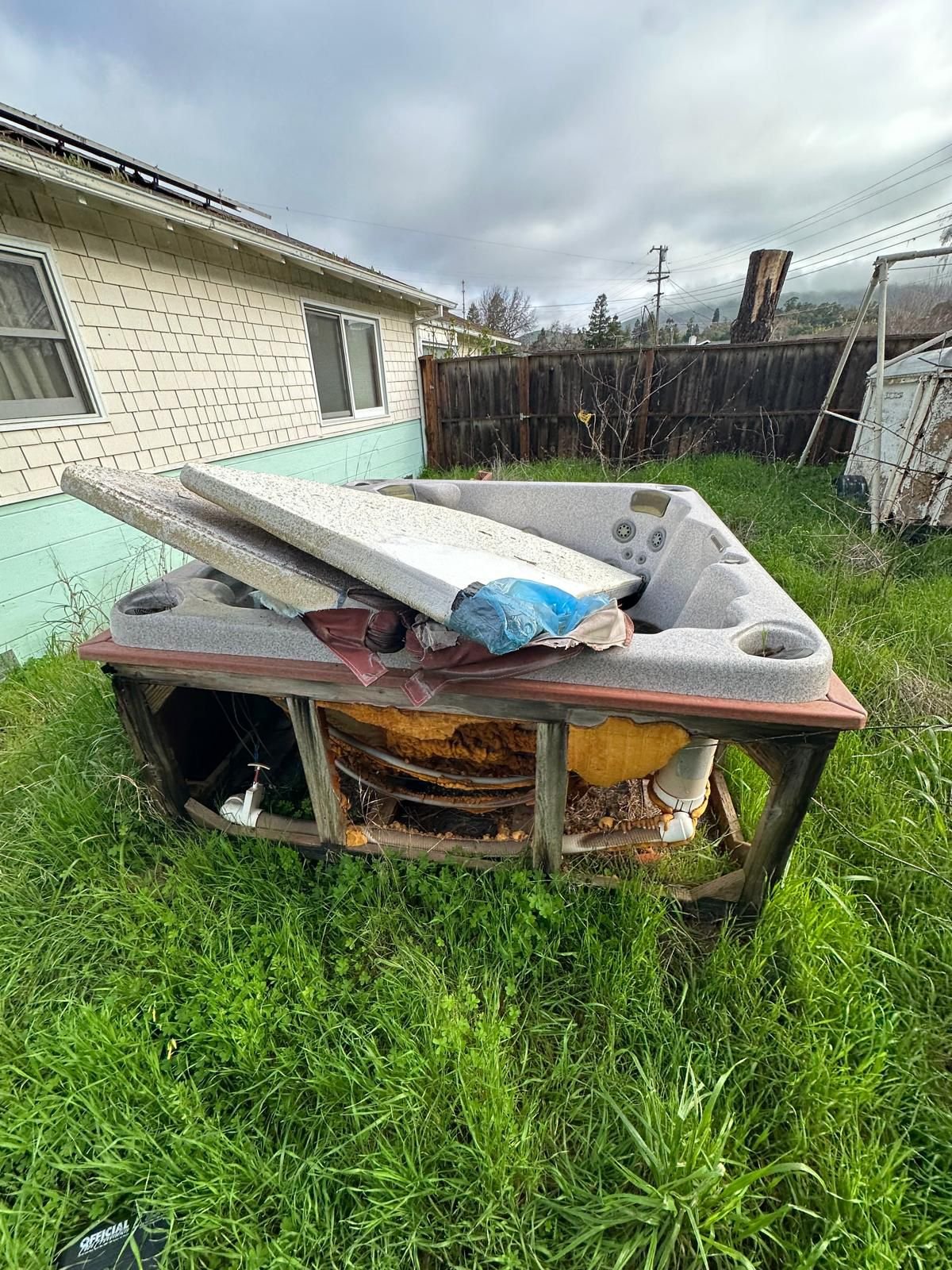 Disassembled hot tub with torn wood frame and overgrown grass in backyard, with house and cloudy sky in background.