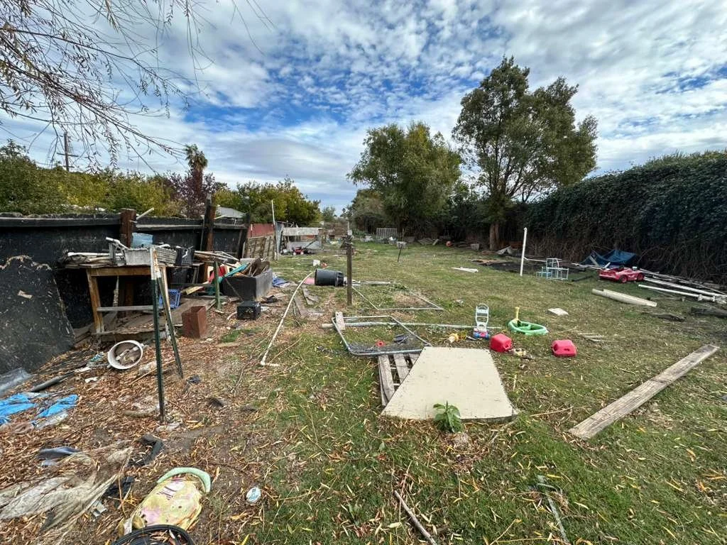 Messy backyard with scattered toys, debris, and uneven grass, enclosed by a fence and trees.
