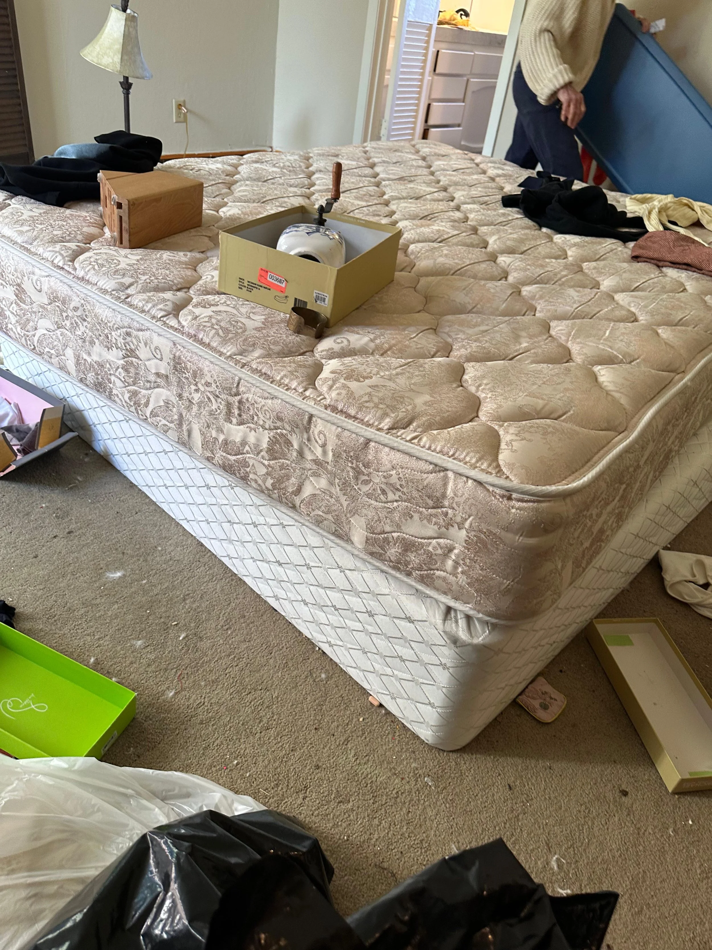 A bedroom with a mattress on the floor, surrounded by various items including a green box, a black trash bag, and miscellaneous objects, with a person standing at the doorway.