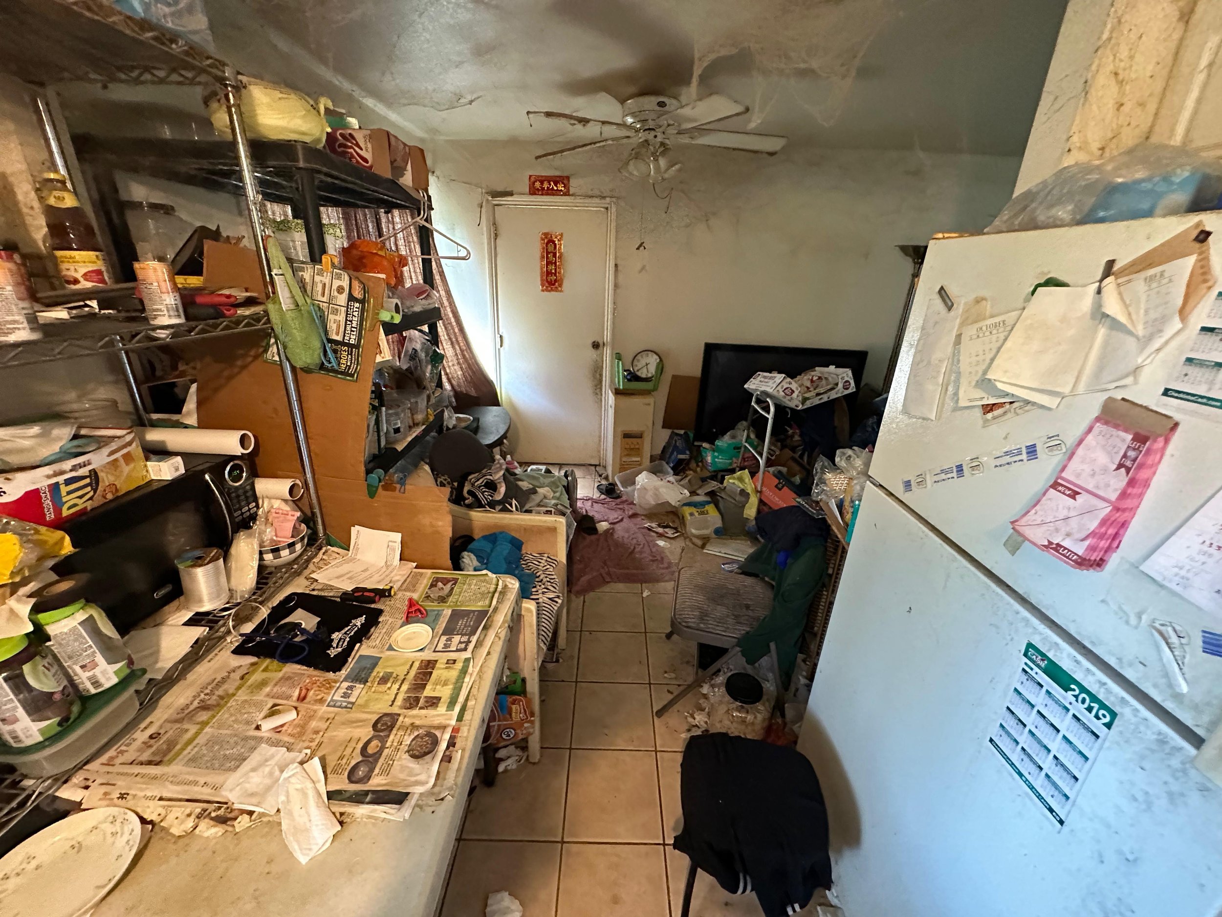 A cluttered and messy kitchen with a table covered in newspapers, dishes, and various items, shelves with cans and boxes, a refrigerator with papers stuck on it, a small chair, a television, and a ceiling fan, all in a room with peeling walls and a tile floor.