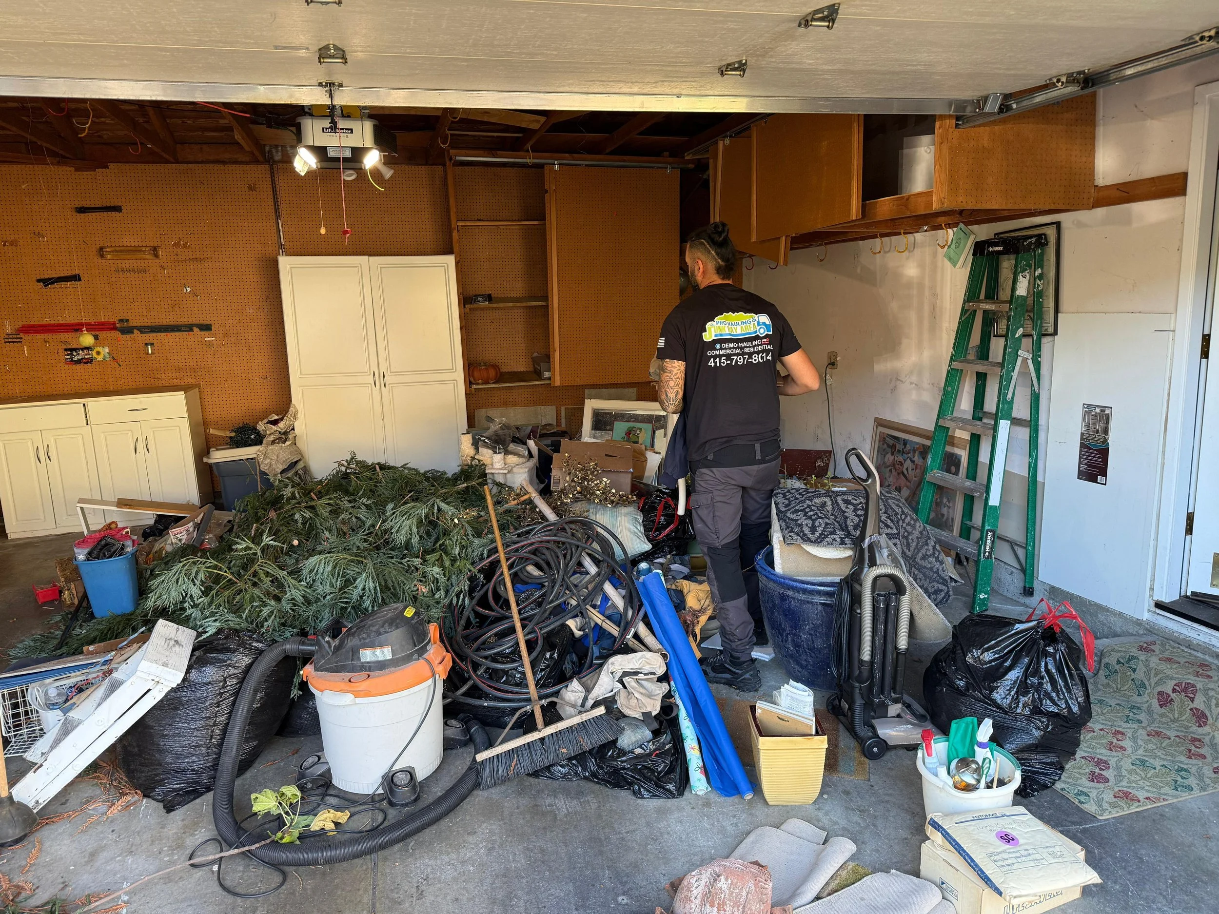 A person in a garage with clutter, including a vacuum, a ladder, bags, and Christmas decorations.