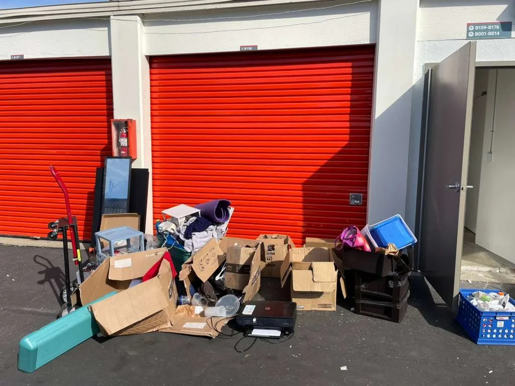 Various discarded items, including cardboard boxes, plastic containers, a small blue table, clothes, and electronic equipment, are piled outside a storage unit with a red rolling door.