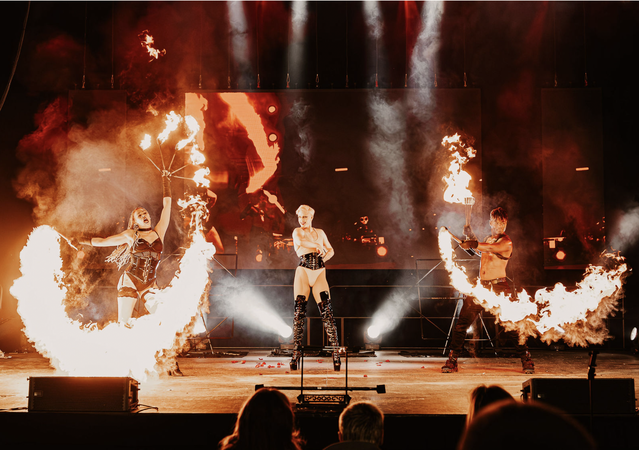 Three performers on stage with fire props, two women with fire rings and a man with a fire torch, during a fire dancing act with a backdrop of flames and bright lights.