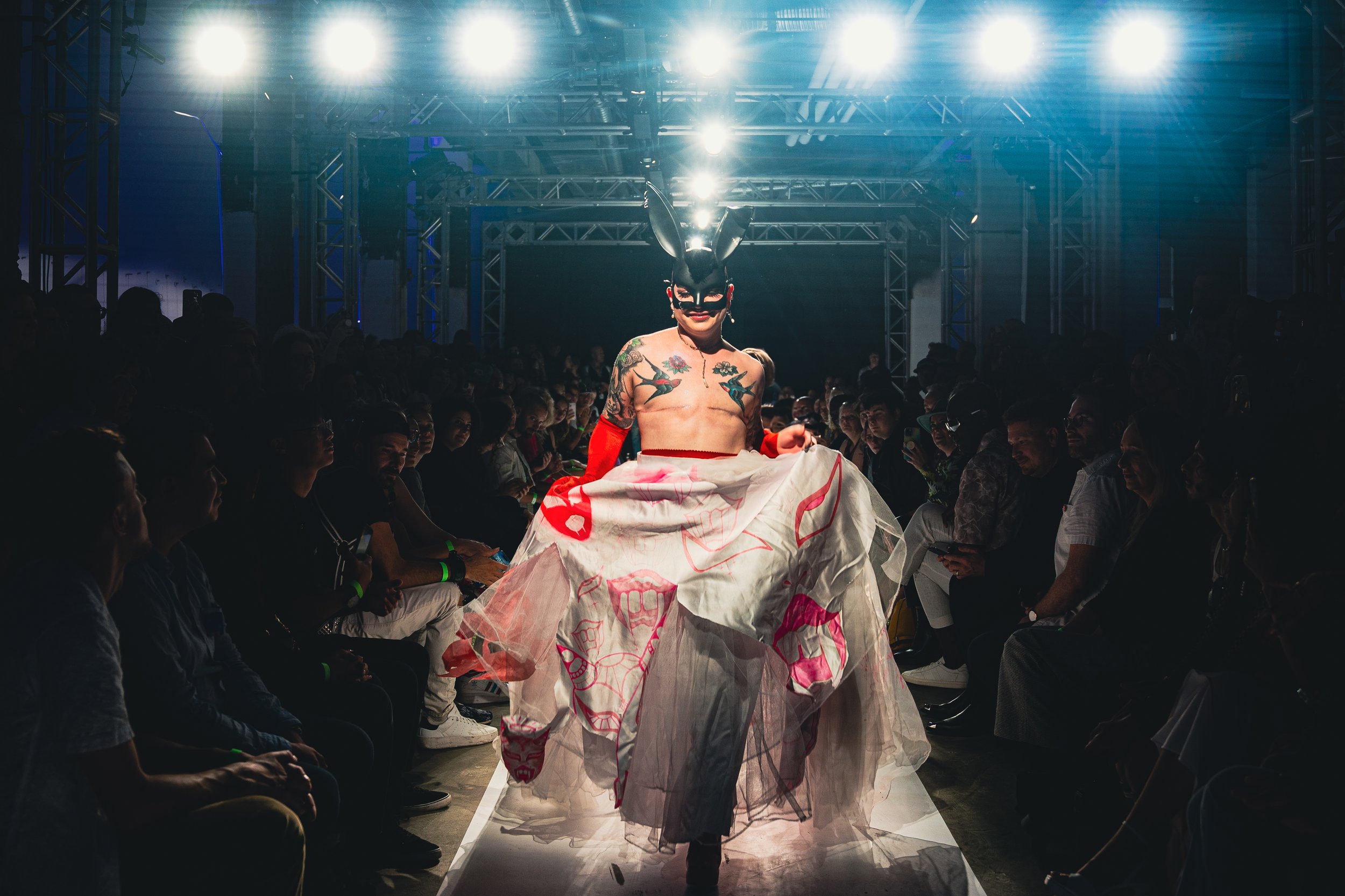 Person walking on runway at fashion show wearing a bunny mask, a sheet with lipstick kiss marks, and gloves, with an audience watching.