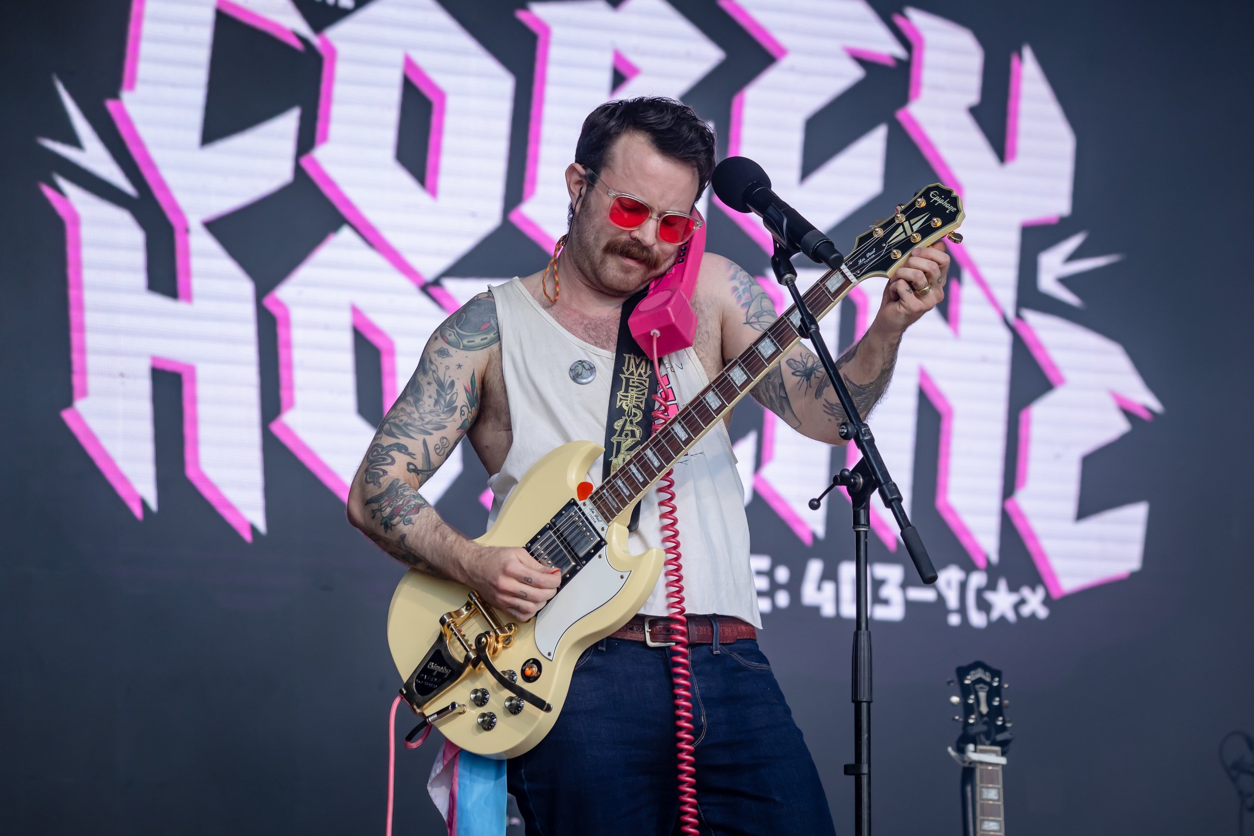 A man with tattoos, red sunglasses, and a mustache is playing an electric guitar on stage, speaking into a pink telephone receiver held between his ear and shoulder. He is standing in front of a large screen with pink and white geometric text.