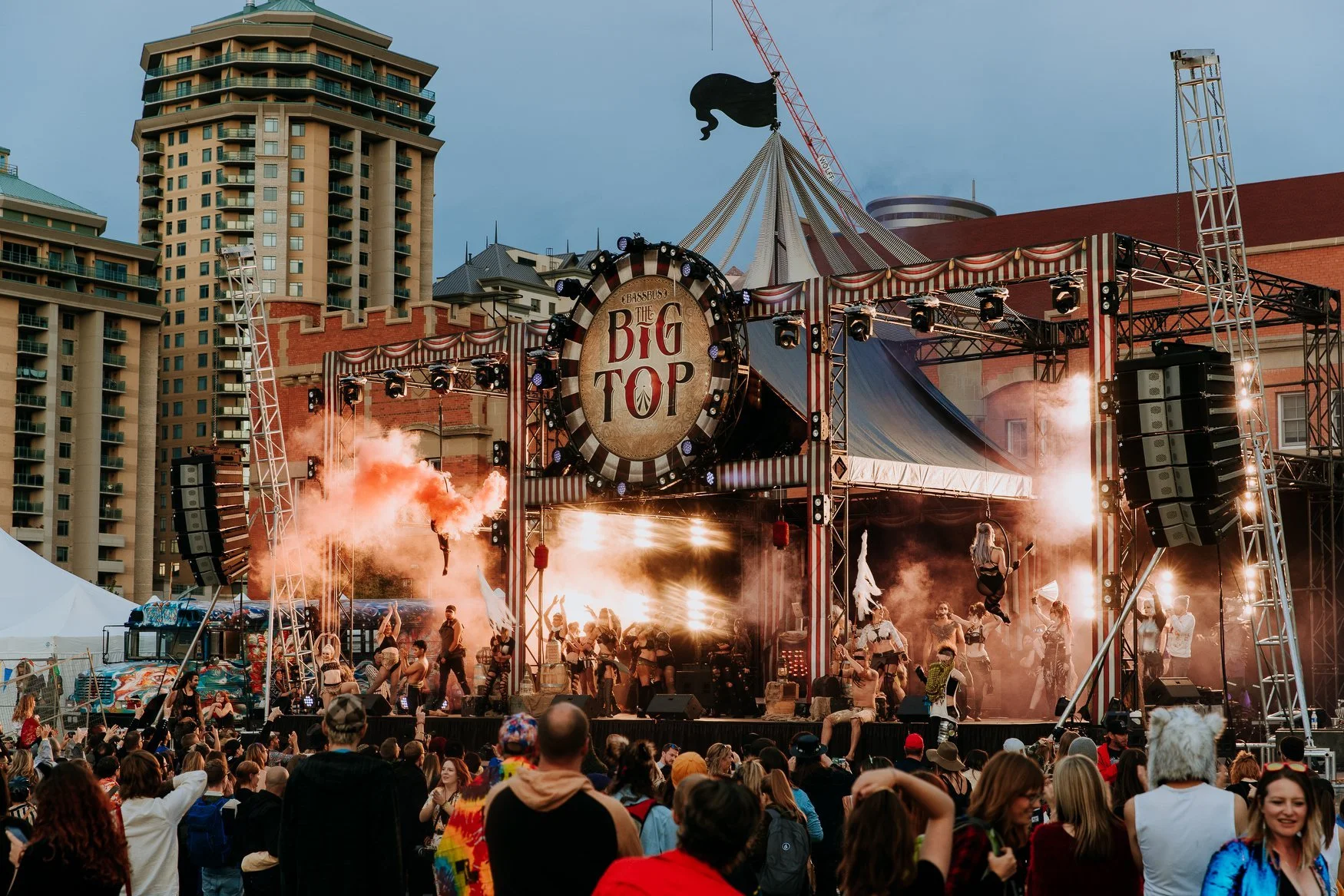 Crowd watching a lively outdoor music concert with performers dancing on stage, large speakers, smoke effects, and a circus-style tent with a sign that says 'Big Top' in an urban setting with tall buildings in the background.