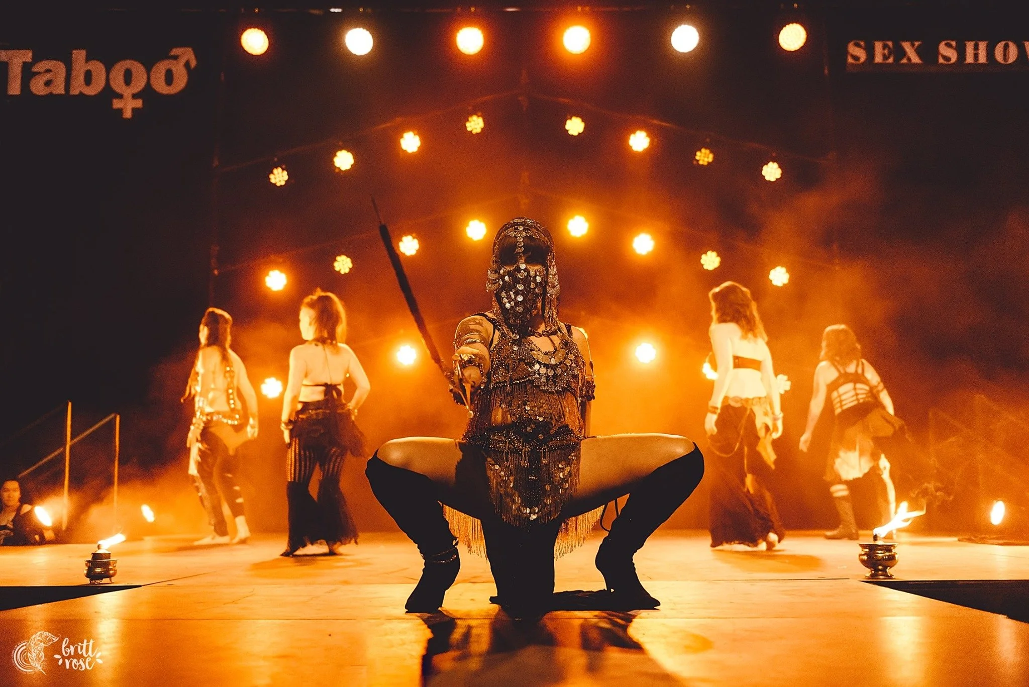 A performer dressed in ornate, black and gold costume kneels in the foreground, holding a spear, with four dancers in similar costumes dancing behind them on stage illuminated by warm orange lights. Signs reading 'Taboo' and 'Sex Show' are visible at