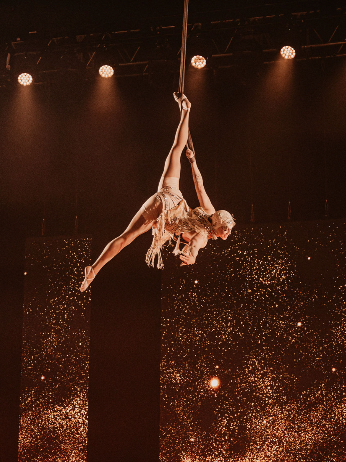 Aerial performer hanging upside down in a star shape during a stage show, with a backdrop of sparkling lights and a dark background.