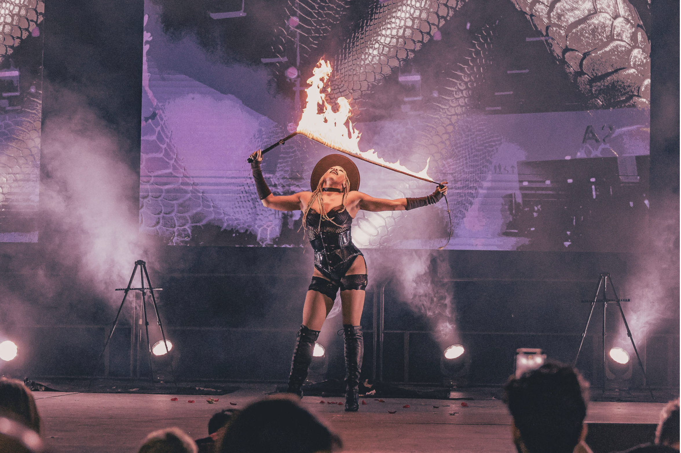 A female performer in a black outfit, black hat, and thigh-high boots holds a flaming torch while standing on stage, with a backdrop of a dragon image and stage lights playing.