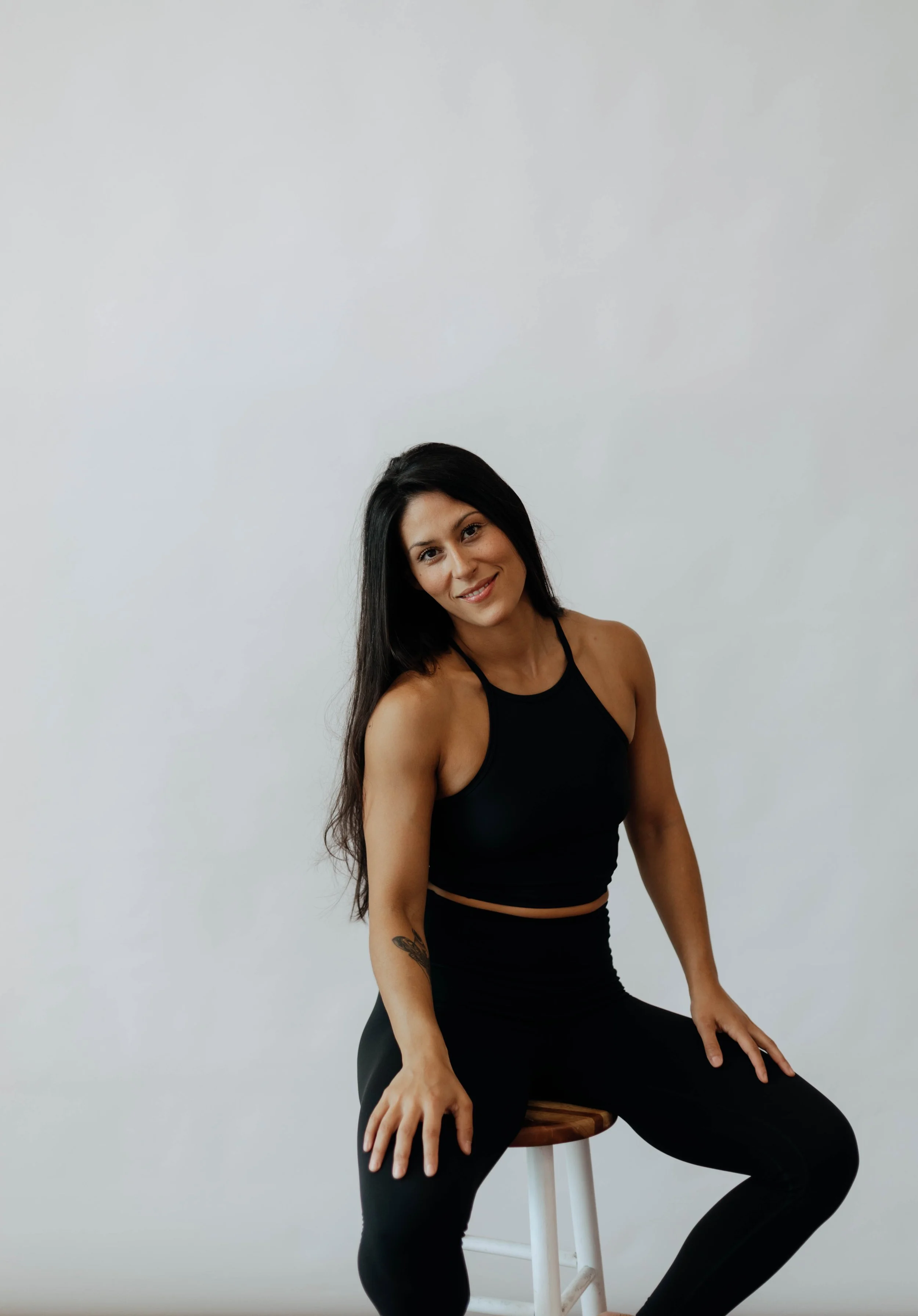 A woman with long black hair sitting on a wooden stool against a plain white background, wearing a black sports top and black leggings, smiling at the camera.