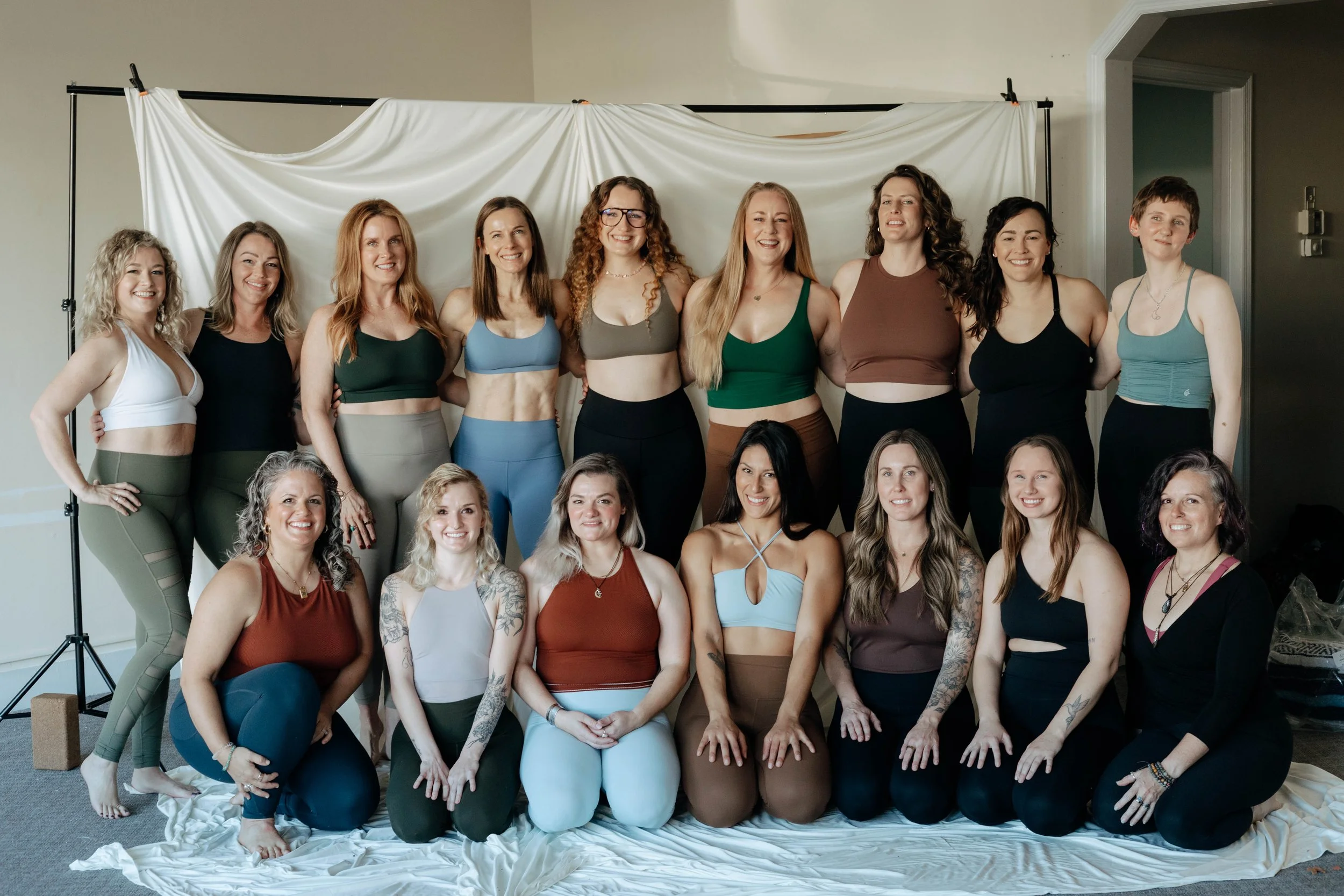 A group of 16 women in athletic wear posing indoors in front of a white backdrop.