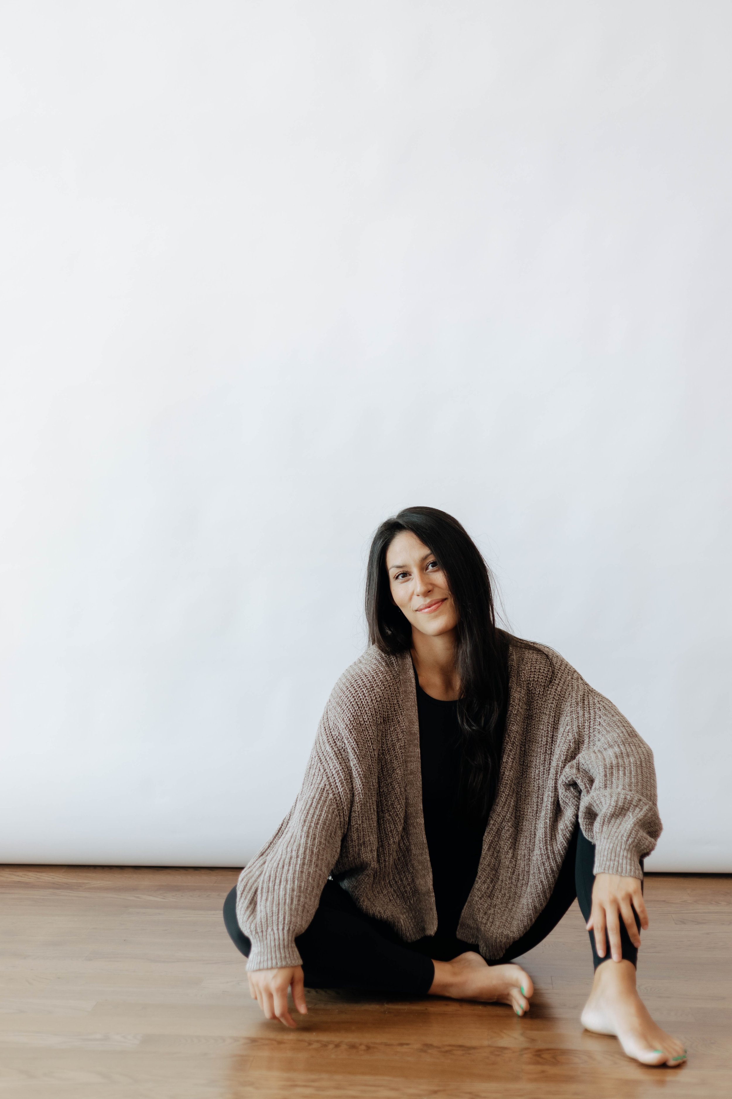 A woman sitting barefoot on a wooden floor against a plain white background, wearing a brown sweater and black pants, smiling at the camera.