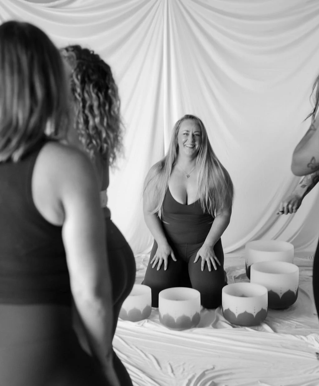 A woman with long blonde hair smiling and kneeling in front of a draped backdrop, surrounded by large singing bowls, with three other women partially visible in the foreground.