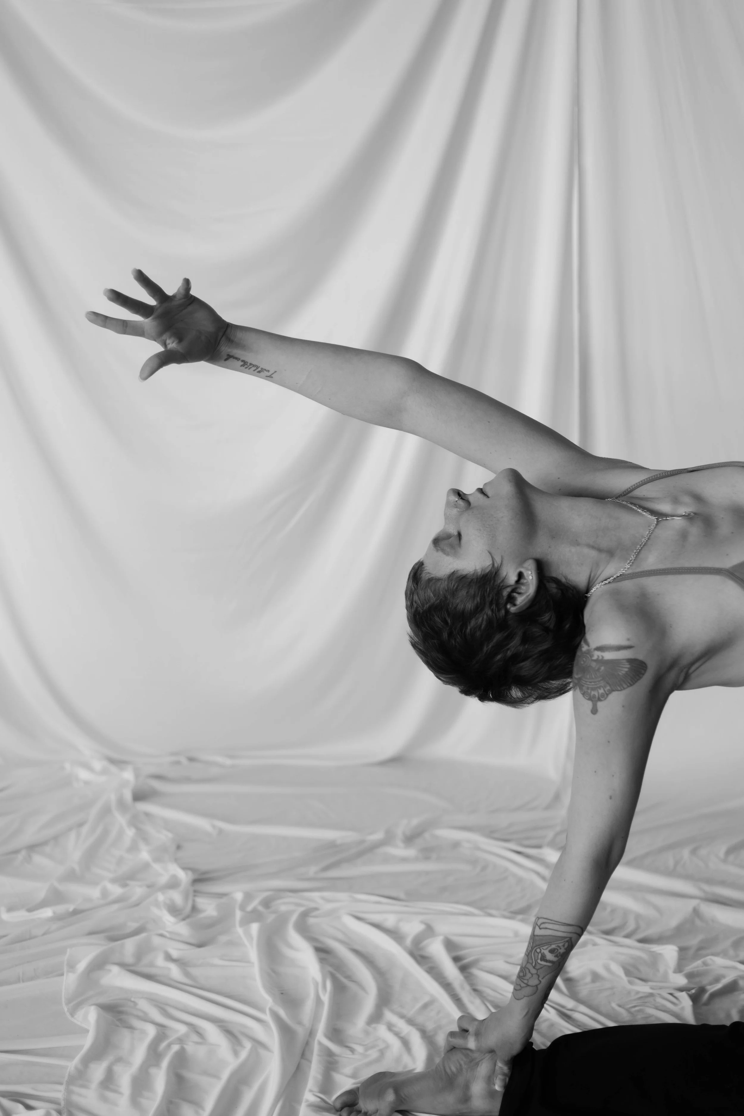 A woman with tattoos on her arms and shoulder, wearing a thin strap top, is performing a yoga pose on a draped fabric backdrop. She is leaning forward with one arm extended upward and the other arm holding onto her foot.