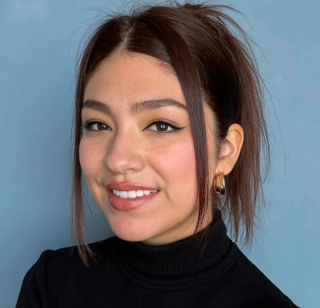A young woman with shoulder-length dark red hair styled with a middle part, wearing a black turtleneck and gold hoop earrings, smiling against a blue background.