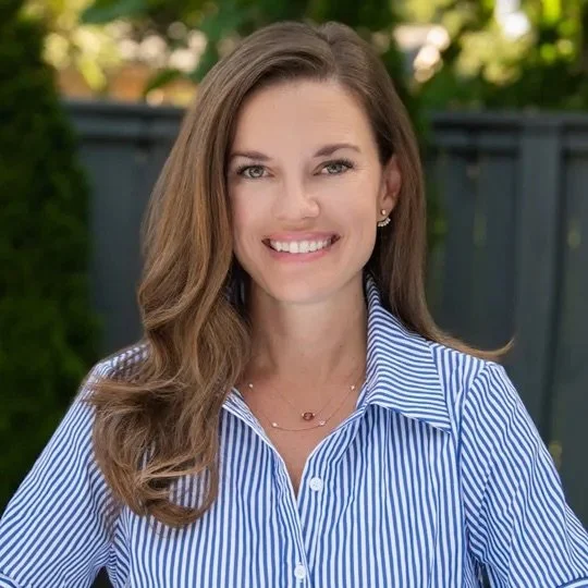 Natalie Warren. A woman with long, wavy brown hair wearing a blue and white striped collared shirt, smiling outdoors with a blurred fence and greenery in the background.