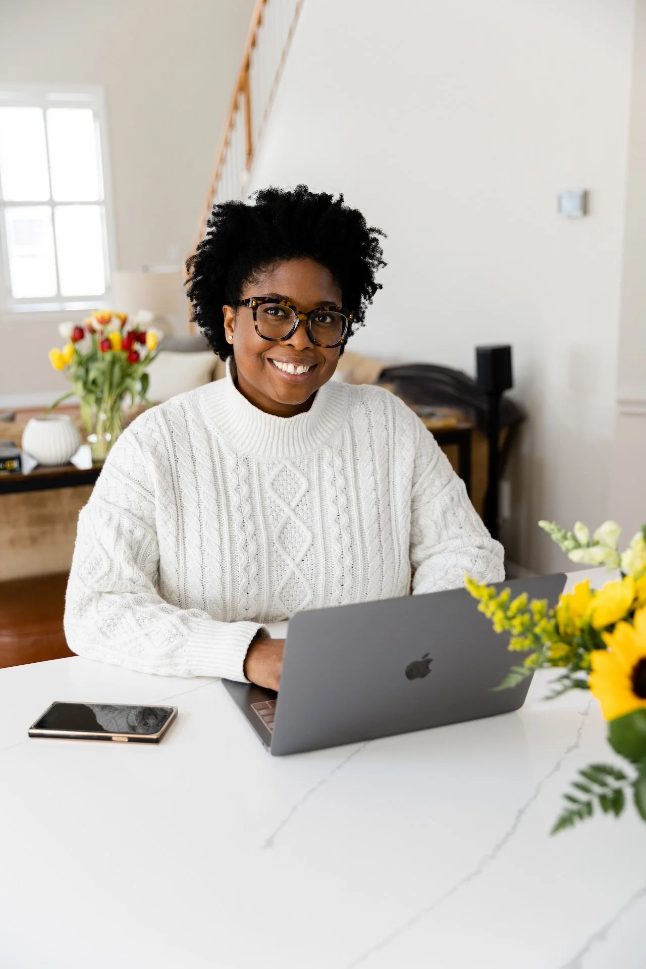 Janica Jordan. A woman with short curly hair, glasses, and a white sweater sitting at a white table with a laptop and smartphone, smiling, in a bright room with flowers on the table and in the background.