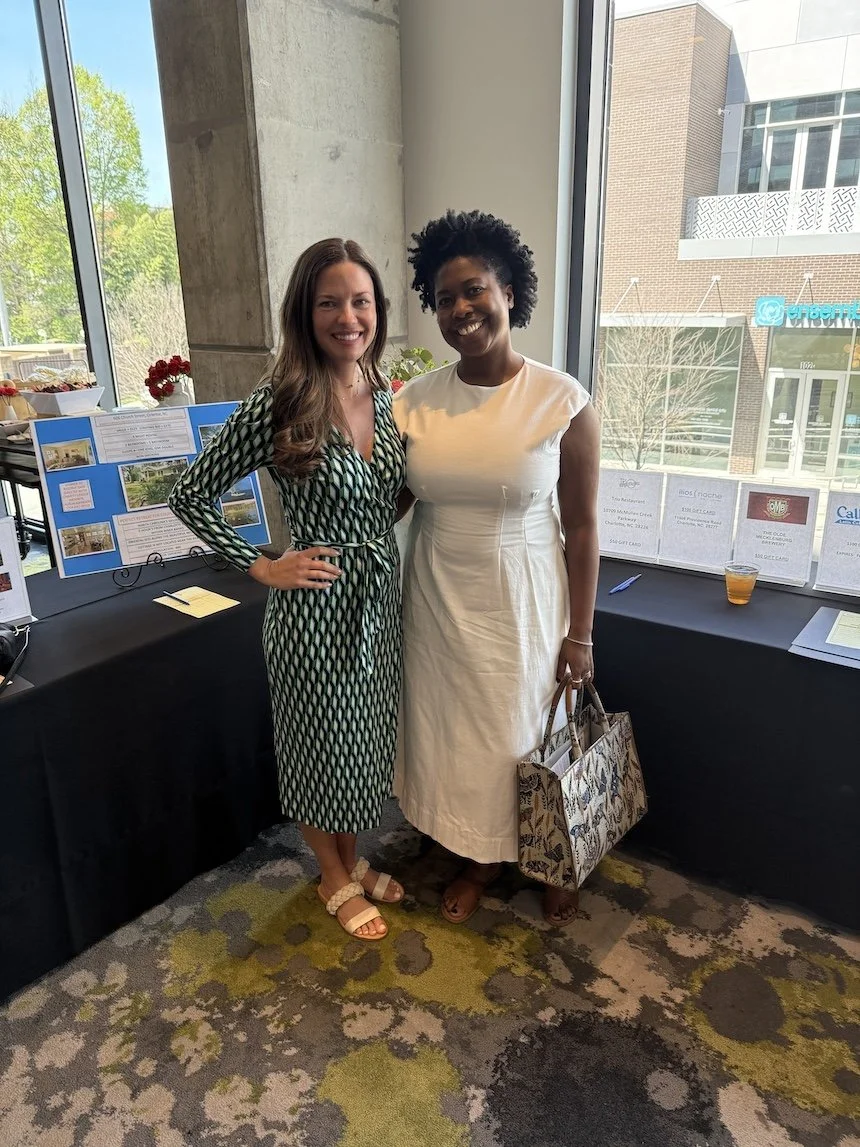 Two women standing side by side inside a building near a large window, smiling at the camera. One woman is wearing a patterned dress, and the other is wearing a white dress with a patterned handbag. There are display boards and drinks on a table behind them.