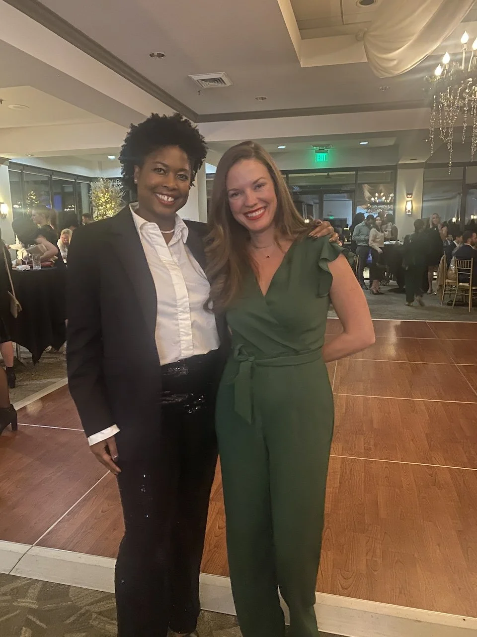 Two women standing together at a formal event, smiling for the camera. One woman is wearing a black suit with a white shirt, and the other is wearing a green jumpsuit. The background shows other people, tables, and elegant decor.