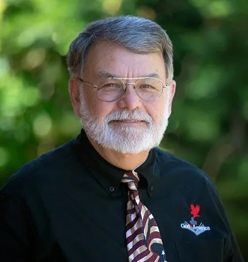A senior man with glasses and a beard, wearing a black shirt with a Canada and Great America logo, standing outdoors with greenery in the background.